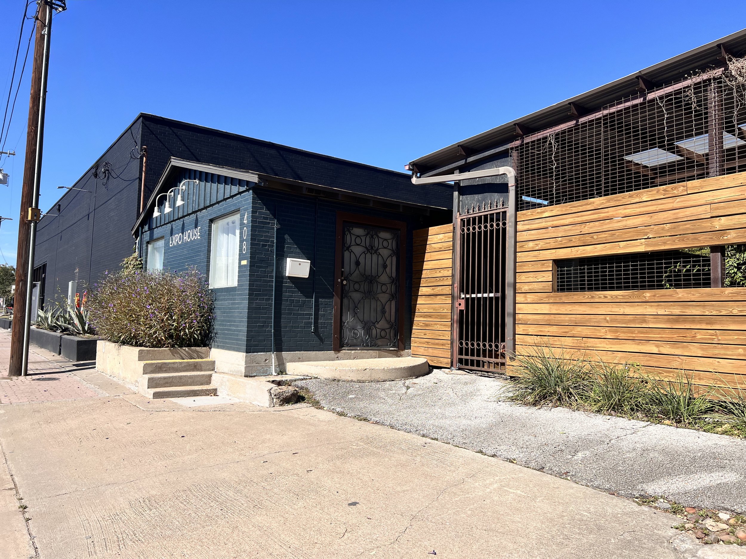 Exterior of a blue brick building with a sign that says 'Expo House', with some plants and sidewalk in front, and a wooden fence and gate on the right side.