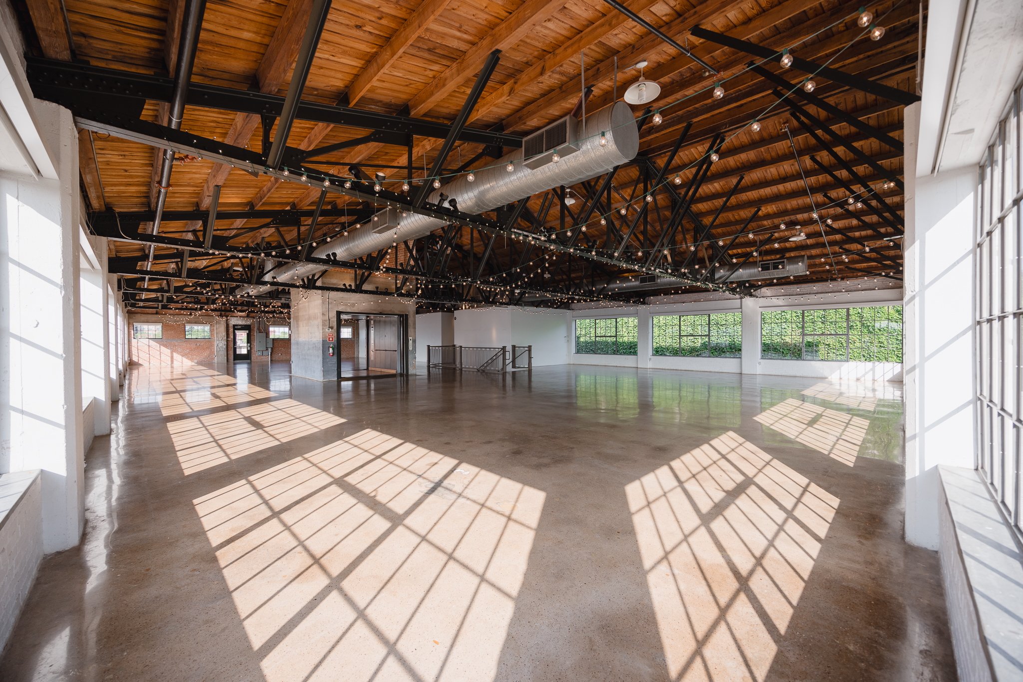Empty industrial-style event space with large windows, polished concrete floor, wooden ceiling with exposed beams, string lights, and outdoor greenery visible through windows.