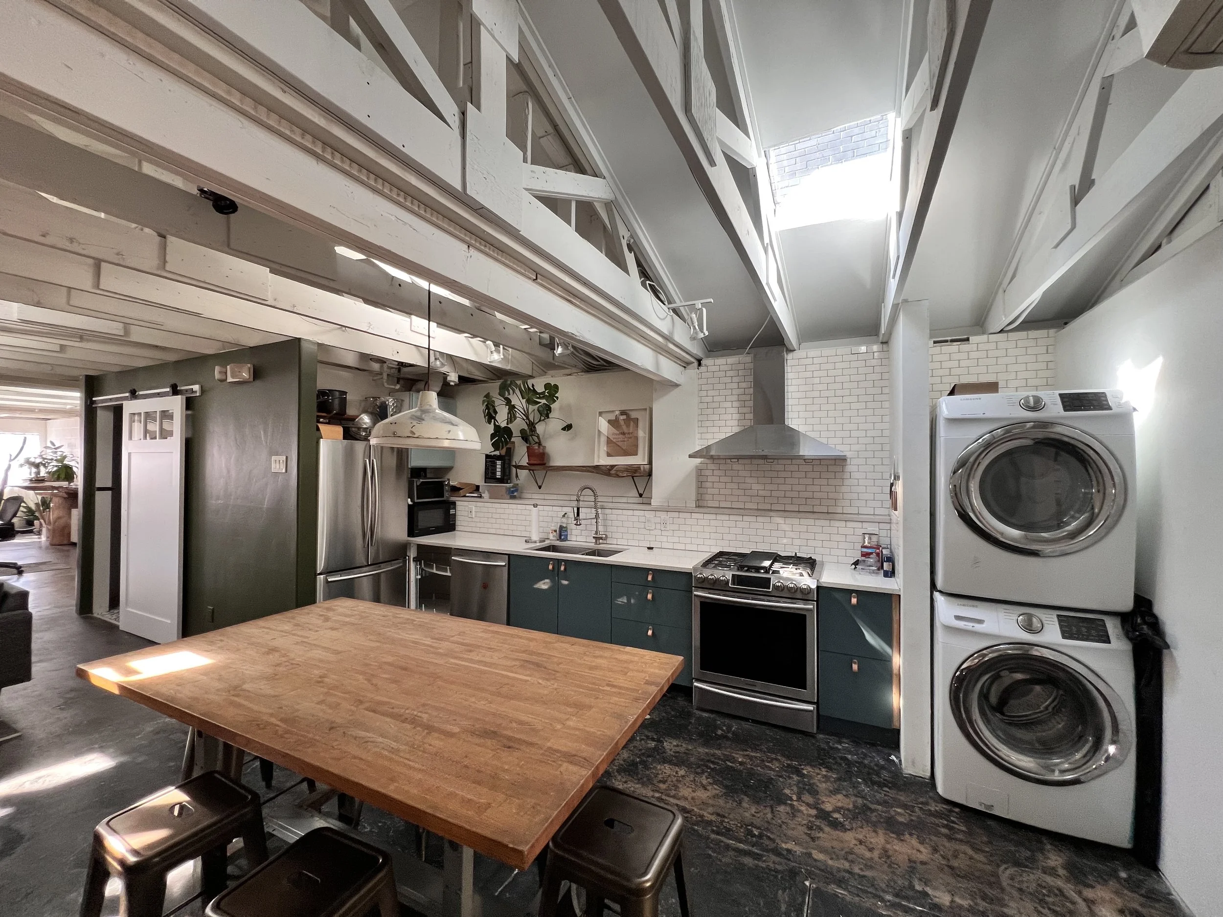 Modern kitchen with a wooden dining table, stacked washer and dryer, kitchen appliances, green cabinets, white subway tile backsplash, and a skylight overhead.