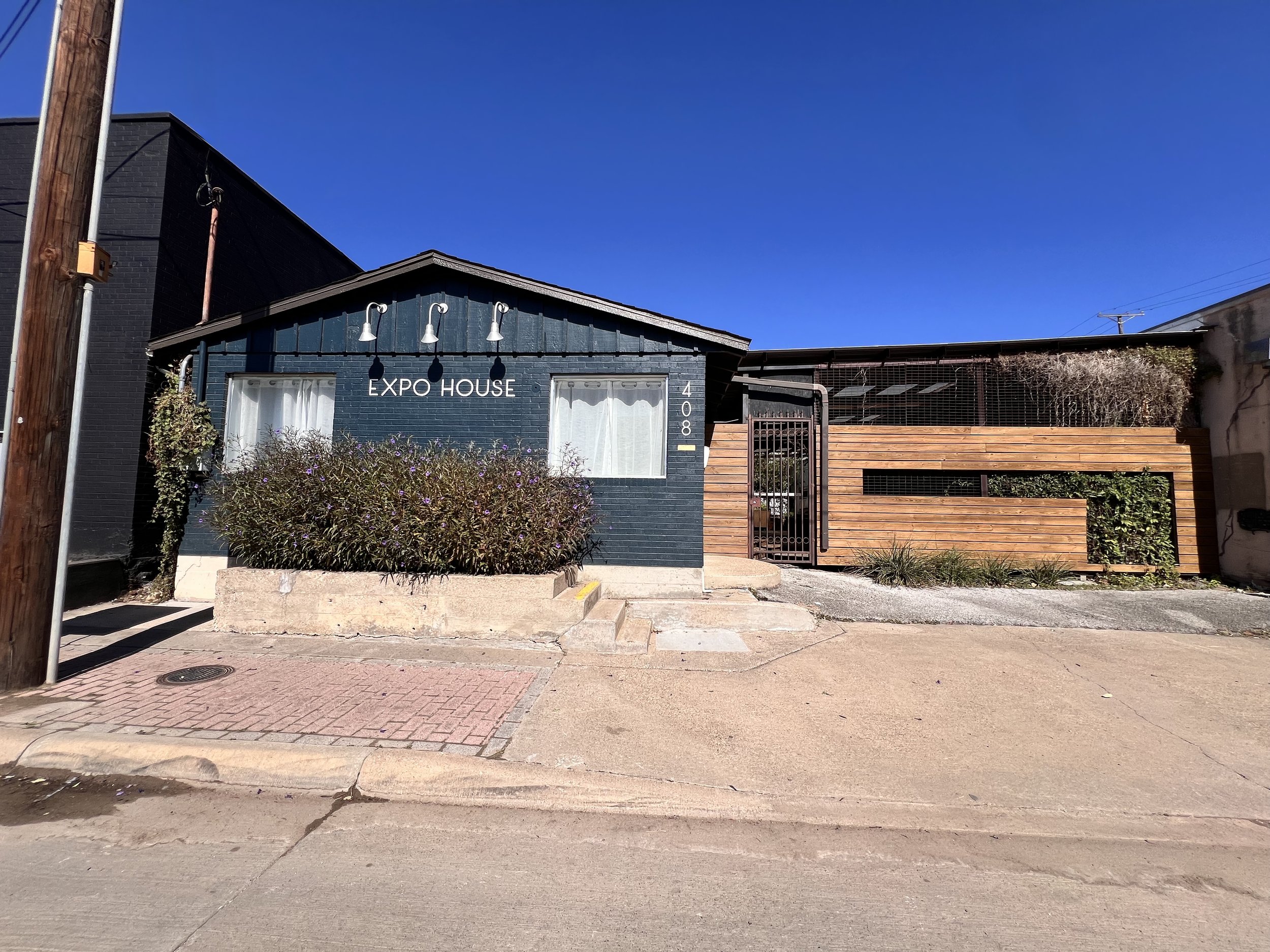 A modern building with the sign 'Expo House' on it, featuring dark blue and wood exterior walls, two windows with white curtains, and a small garden with shrubs in front. The building has address number 408.