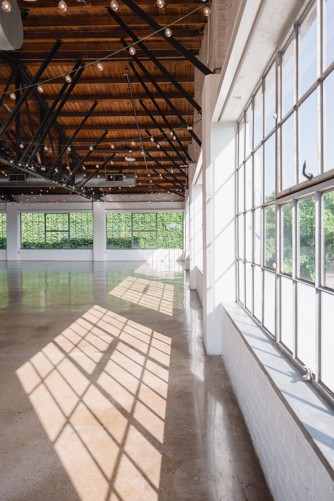 Bright indoor space with large windows letting in sunlight, wooden ceiling beams, string lights, polished concrete floor, and greenery outside.