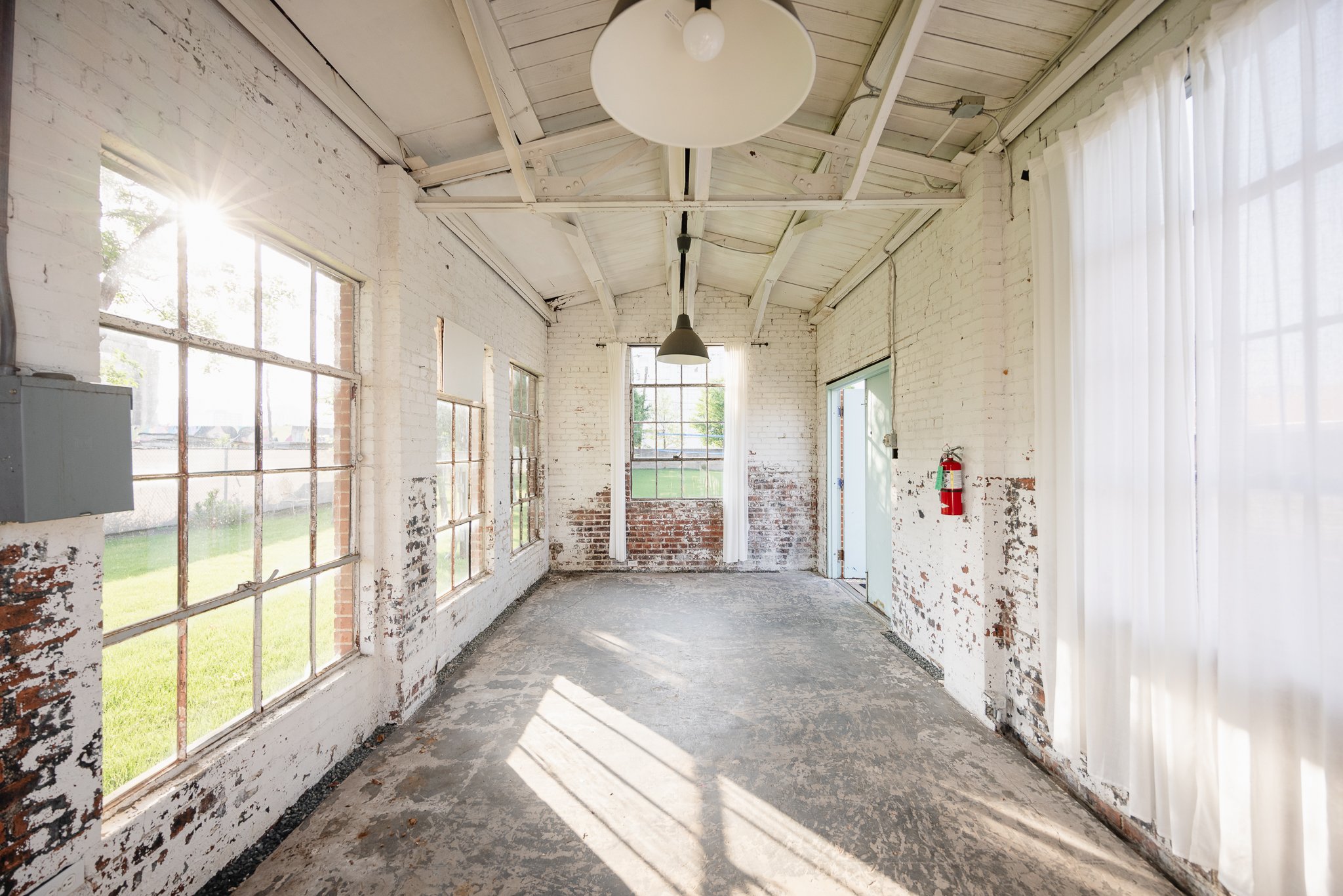 Empty sunlit room with white brick walls, large windows, peeling paint, a fire extinguisher on the right wall, exit door, and a white curtain on the right side.