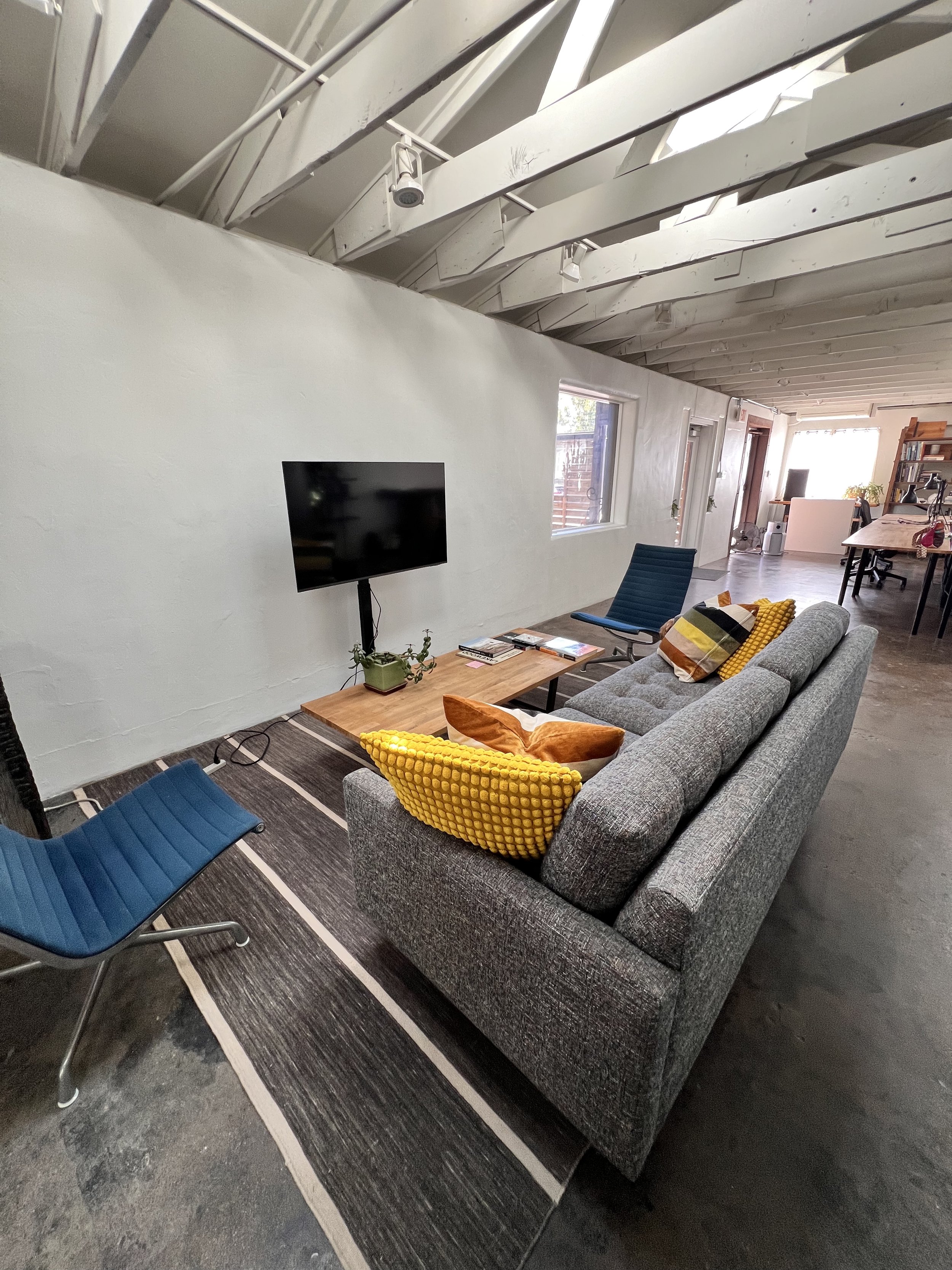 Living room with a gray couch, yellow and brown pillows, a wooden coffee table with magazines, and a blue lounge chair. A flat-screen TV is mounted on a white wall near a window. The ceiling has exposed white beams and track lighting. The floor is da