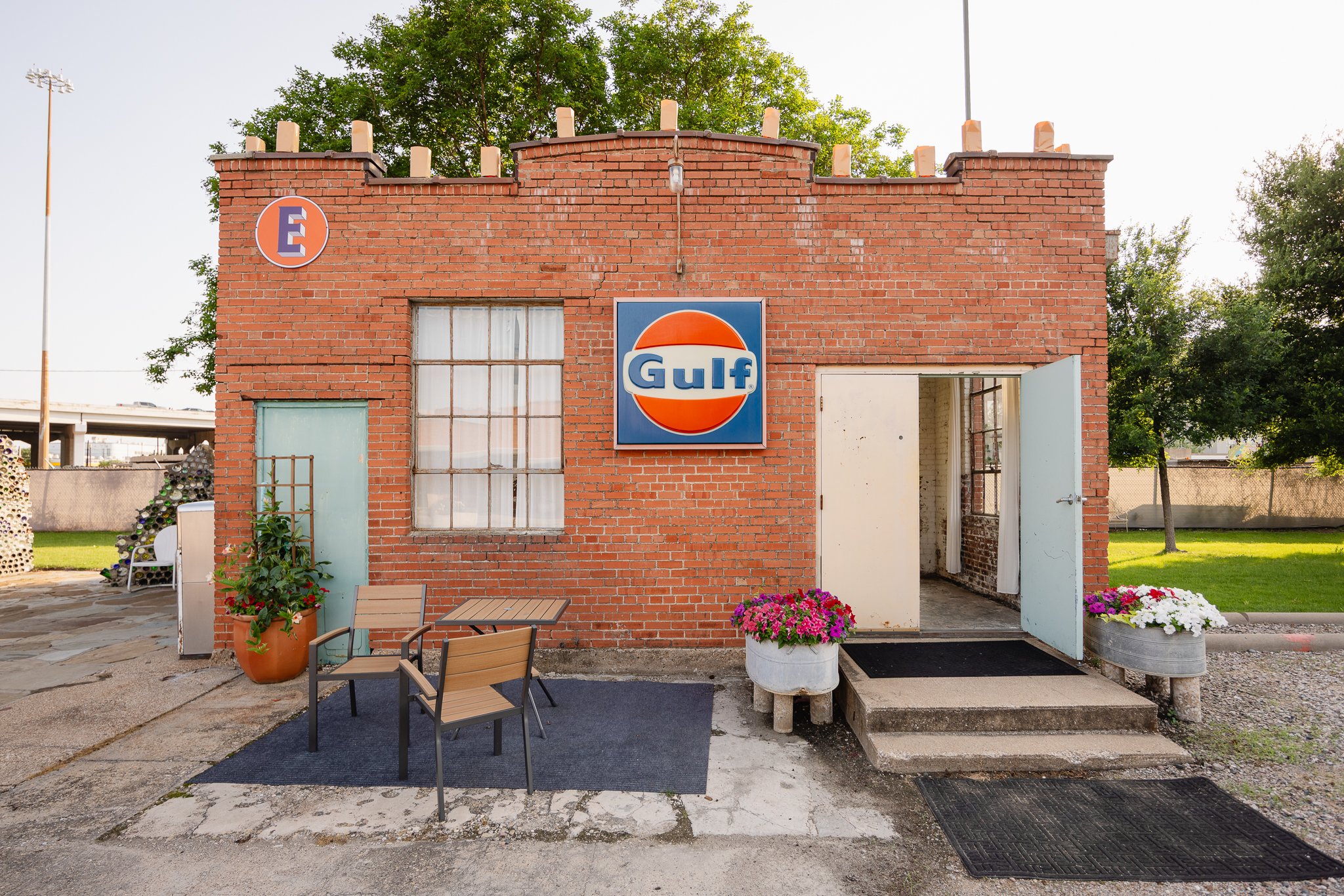A small brick building with an old Gulf gas station sign on the front wall. There are two potted flowers, a table and chairs outside, and a door that is partly open. Green trees and a grassy area are in the background.
