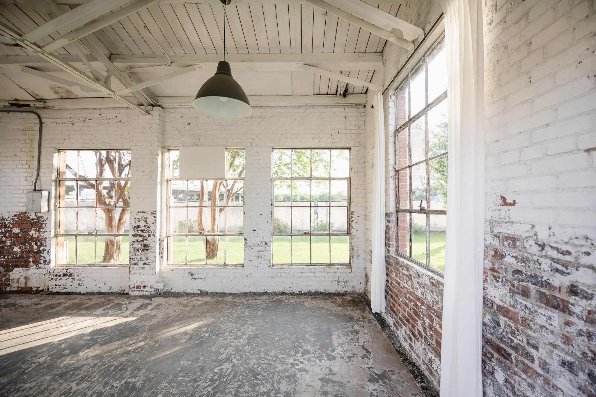 Empty room with large windows, white brick walls, and a concrete floor, sunlight streaming in.