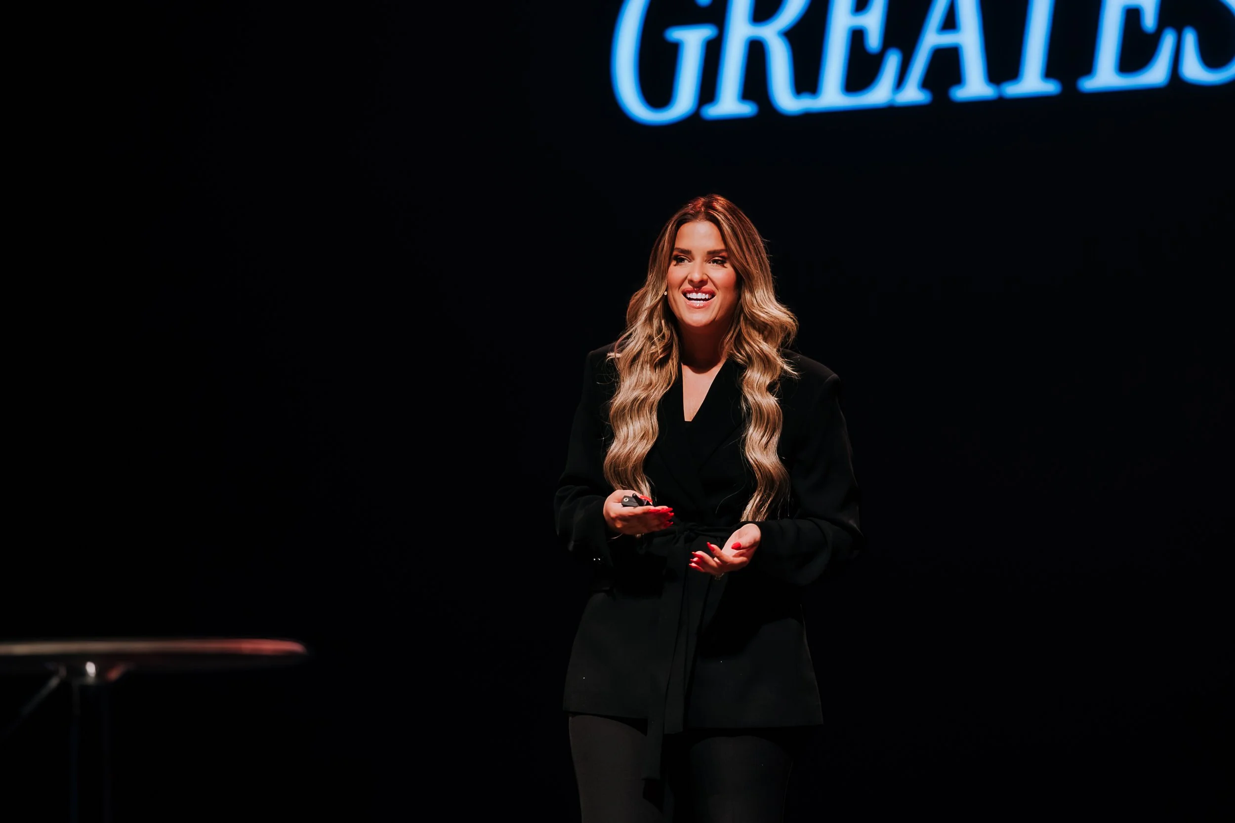 Shannon Gillette wearing a black blazer and holding a remote control, standing in front of a dark background with the illuminated blue word 'GREATNESS' partially visible above her.