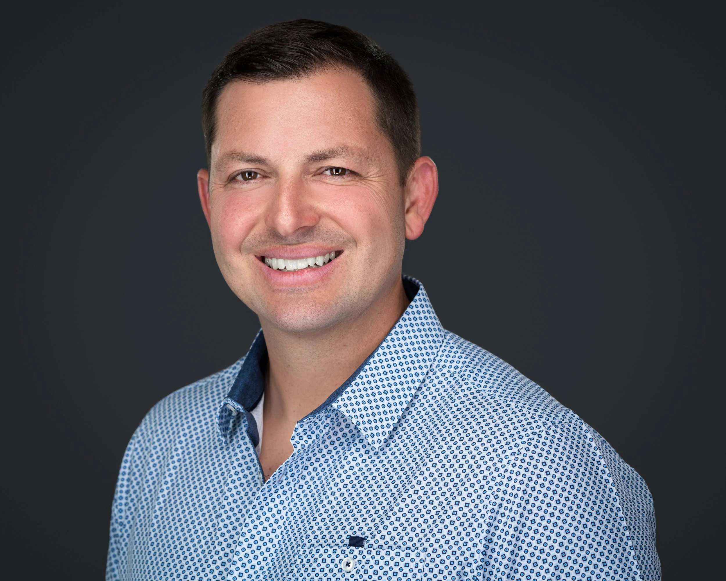 Headshot of a smiling man with short dark hair, wearing a blue patterned dress shirt, against a dark gray background.
