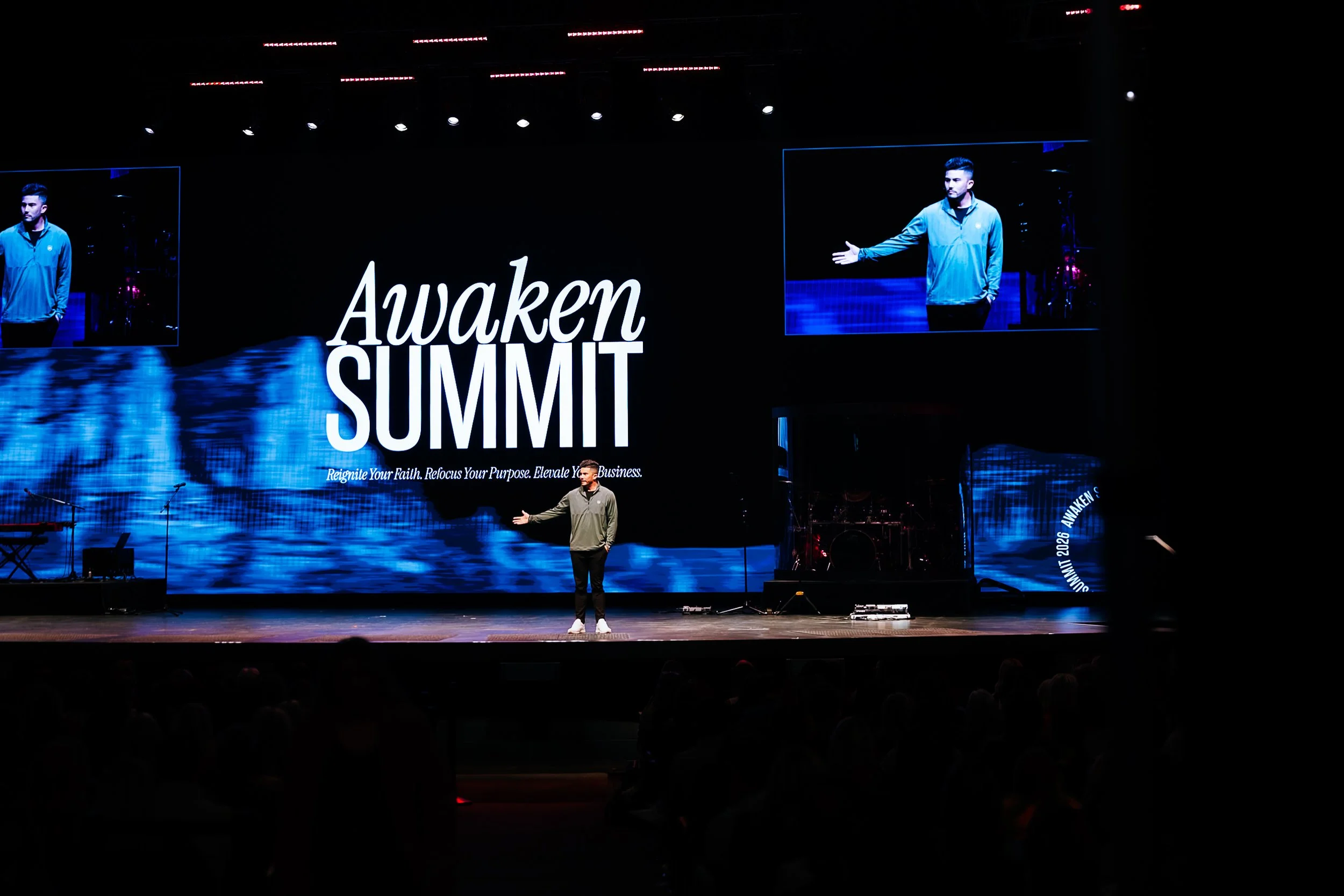 Steve Weatherford, on stage with large screen behind him displaying the words 'Awaken SUMMIT'. The stage is dark with two screens showing a man in a blue jacket gesturing with his right hand. Audience silhouettes are visible in the foreground.