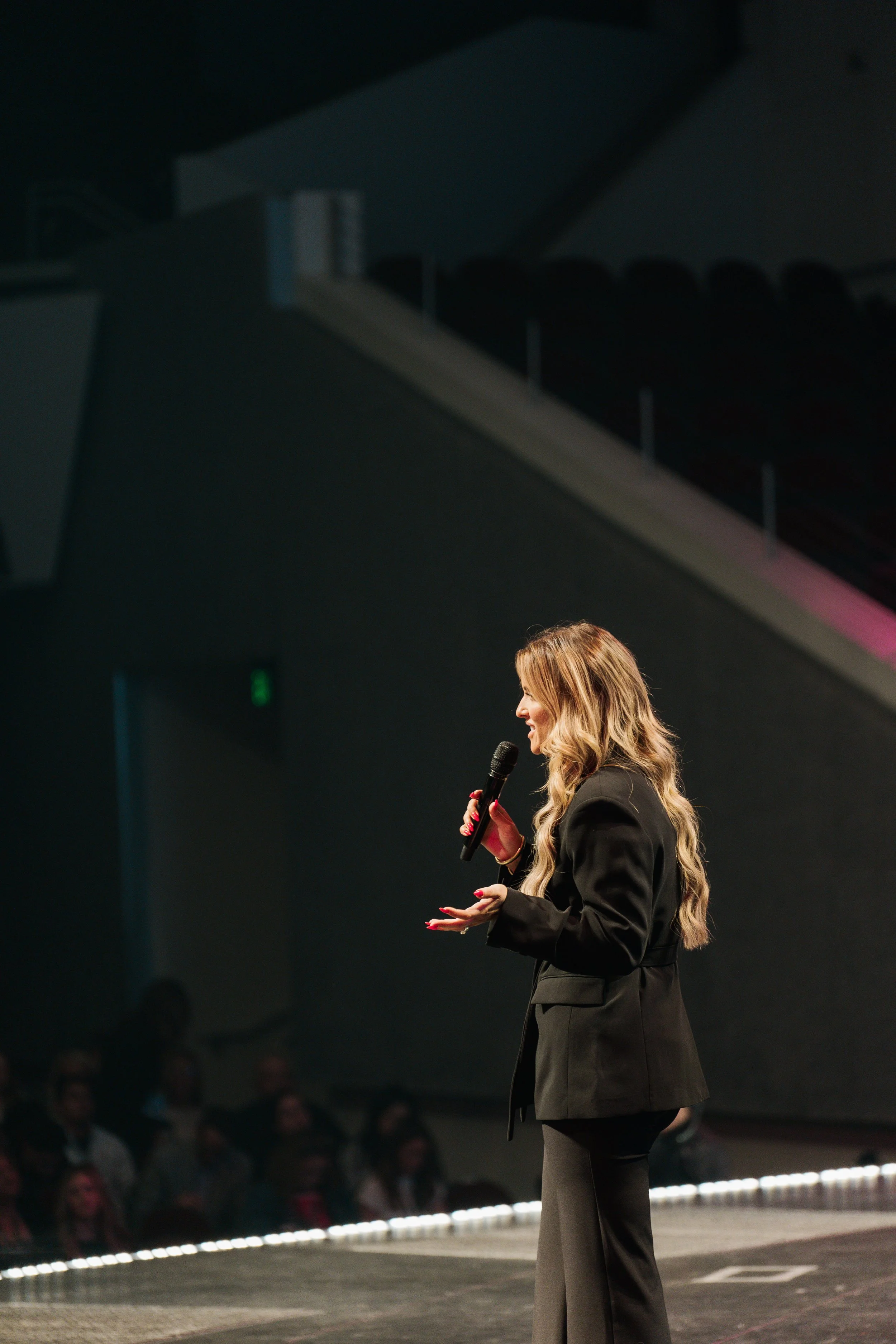 Shannon Gillette, dressed in a black blazer and pants, speaking into a microphone on stage at a conference or event, with an audience seated in front of her.