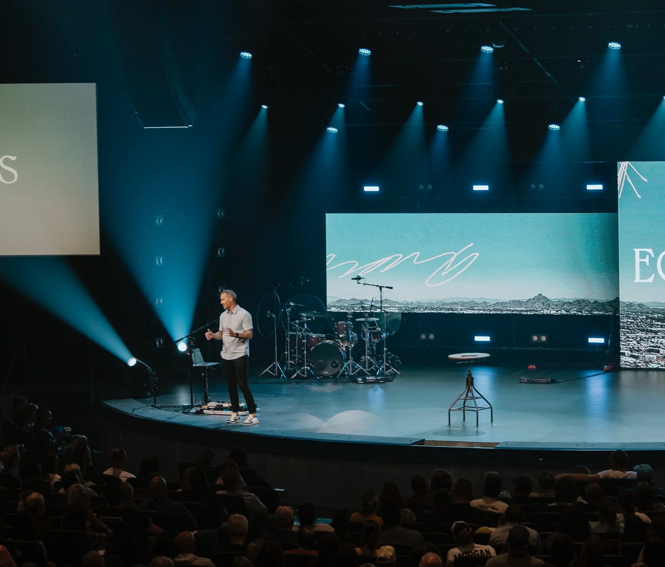 A man is giving a presentation on stage in front of an audience, with a large digital screen displaying graphics behind him. The stage is equipped with musical instruments and lighting.