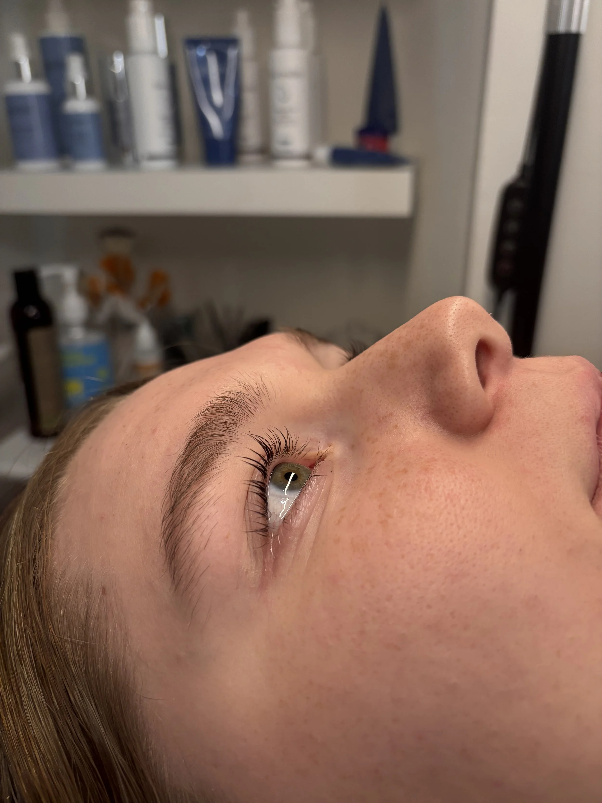Close-up of a woman's face lying down, showing her eye, eyebrow, and nose with a blurred background of skincare products on a shelf.