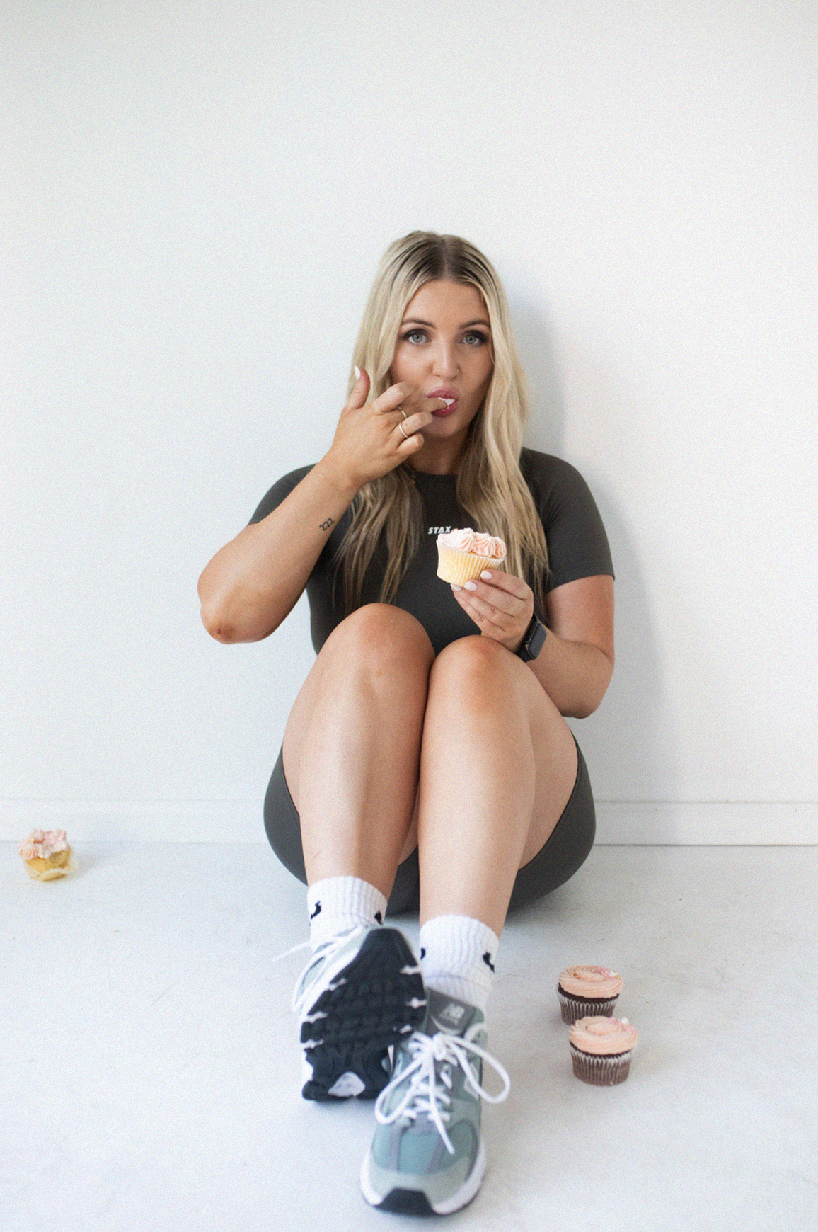 Jess Morgan sitting on the floor against a white wall, holding a cupcake and eating it, with other cupcakes on the floor around her.