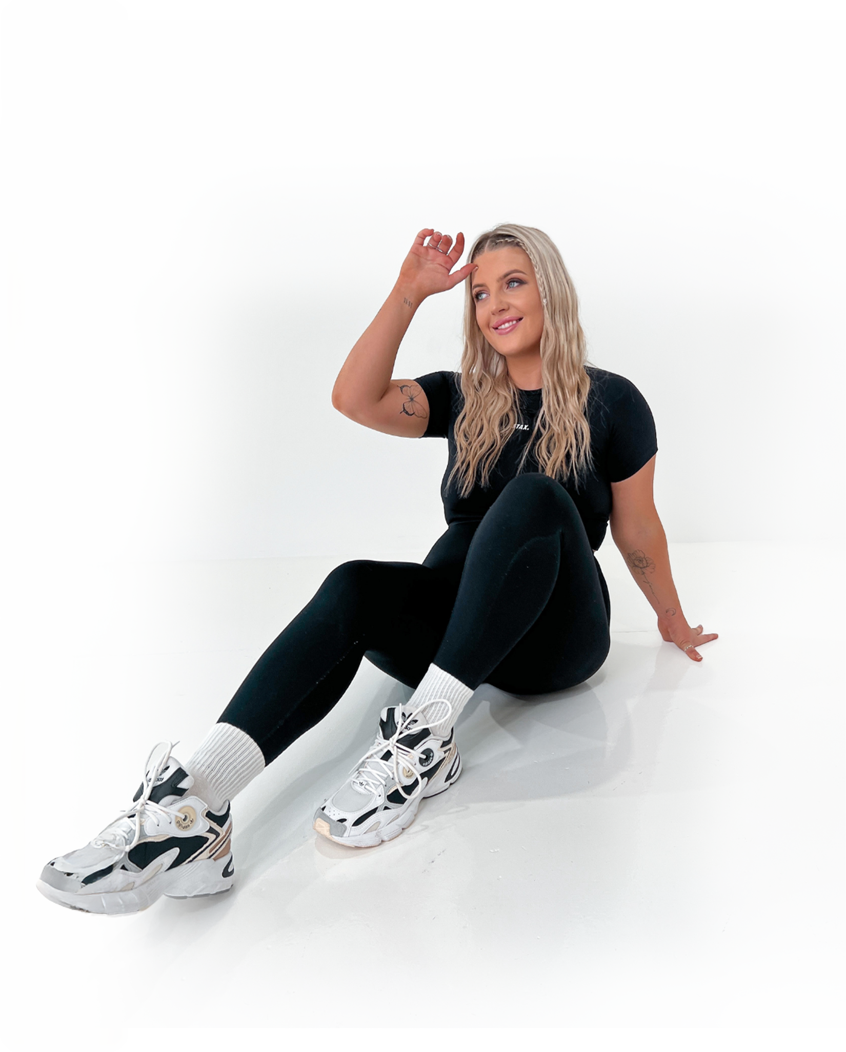 Jess Morgan sitting on the floor against a plain white background, wearing a black t-shirt, black leggings, white athletic shoes, and white socks, smiling and holding her hand above her head.