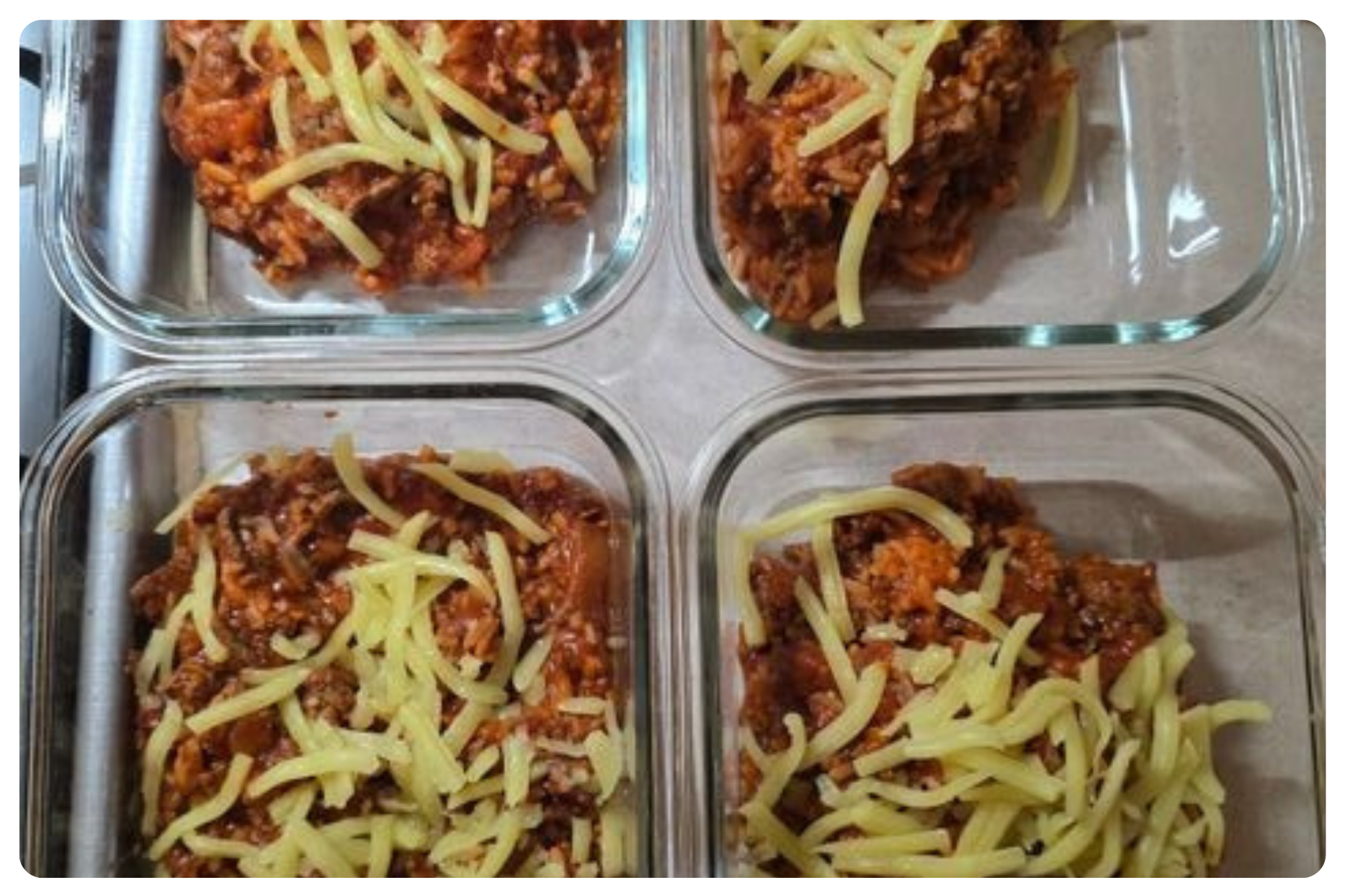 Four glass containers filled with uncooked baked ziti topped with shredded cheese, arranged on a baking sheet.