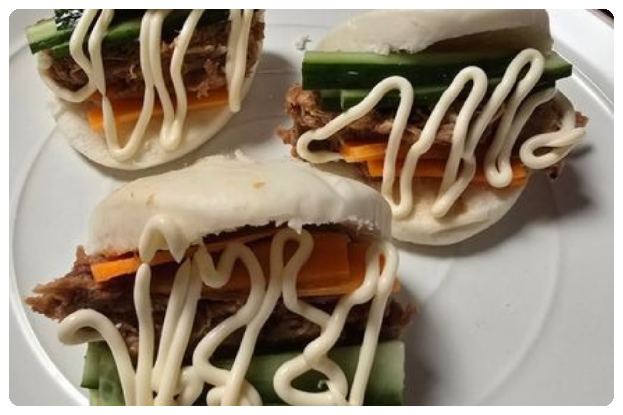 Three homemade steamed bao buns filled with shredded beef, sliced vegetables, and garnished with mayonnaise, on a white plate