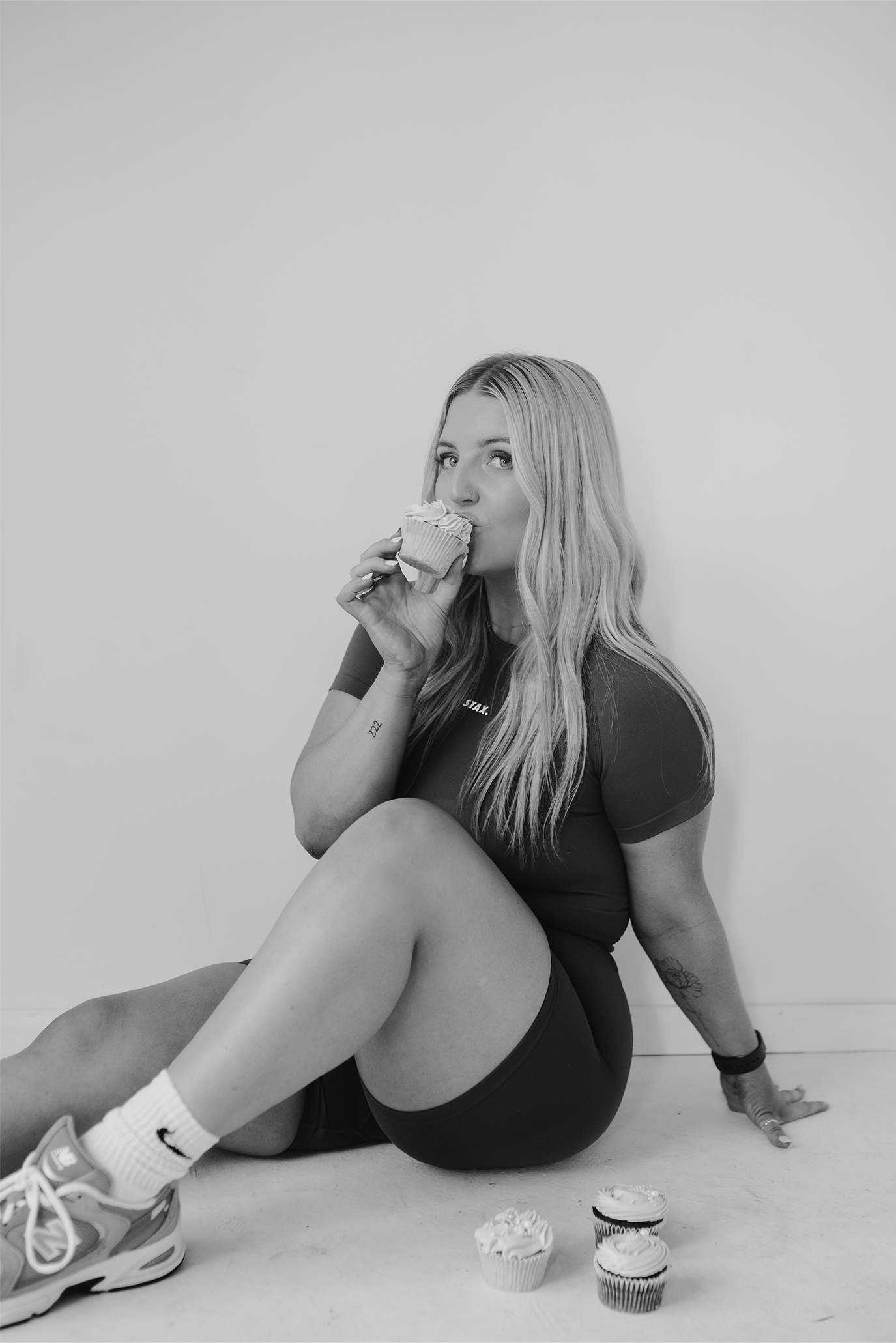 Jess Morgan sitting on the floor against a plain wall, holding a cupcake near her mouth, with three more cupcakes on the floor nearby.