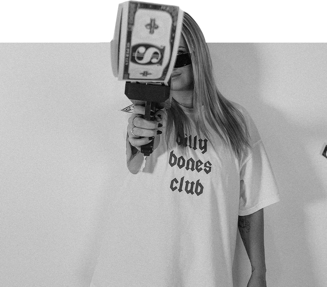Jess Morgan holding a gun-shaped object covered with dollar bill art, wearing a T-shirt that says "dying bones club," with long hair and glasses, standing against a plain wall.