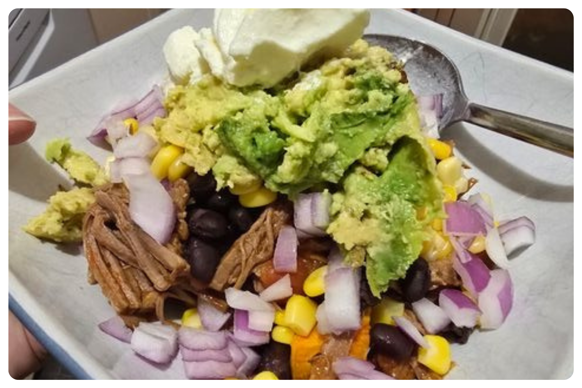 A plate of salad with chopped red onions, black beans, corn, shredded chicken, sliced avocado, a scoop of guacamole, and a dollop of sour cream, on a white surface with a spoon.