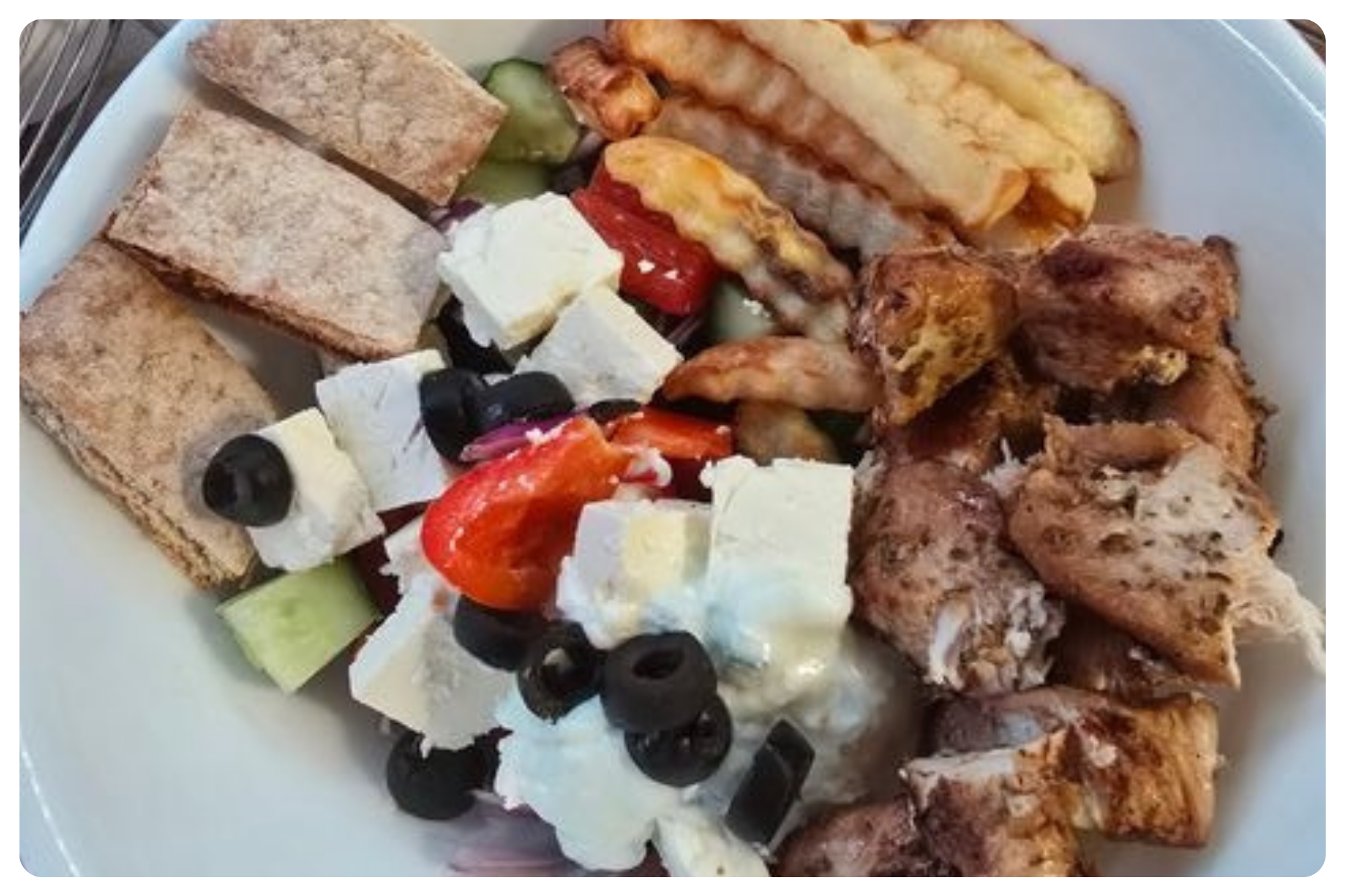 A plate of assorted food including grilled chicken pieces, Greek salad with feta cheese, black olives, cherry tomatoes, cucumber, and peppers, along with slices of bread and fried plantains.