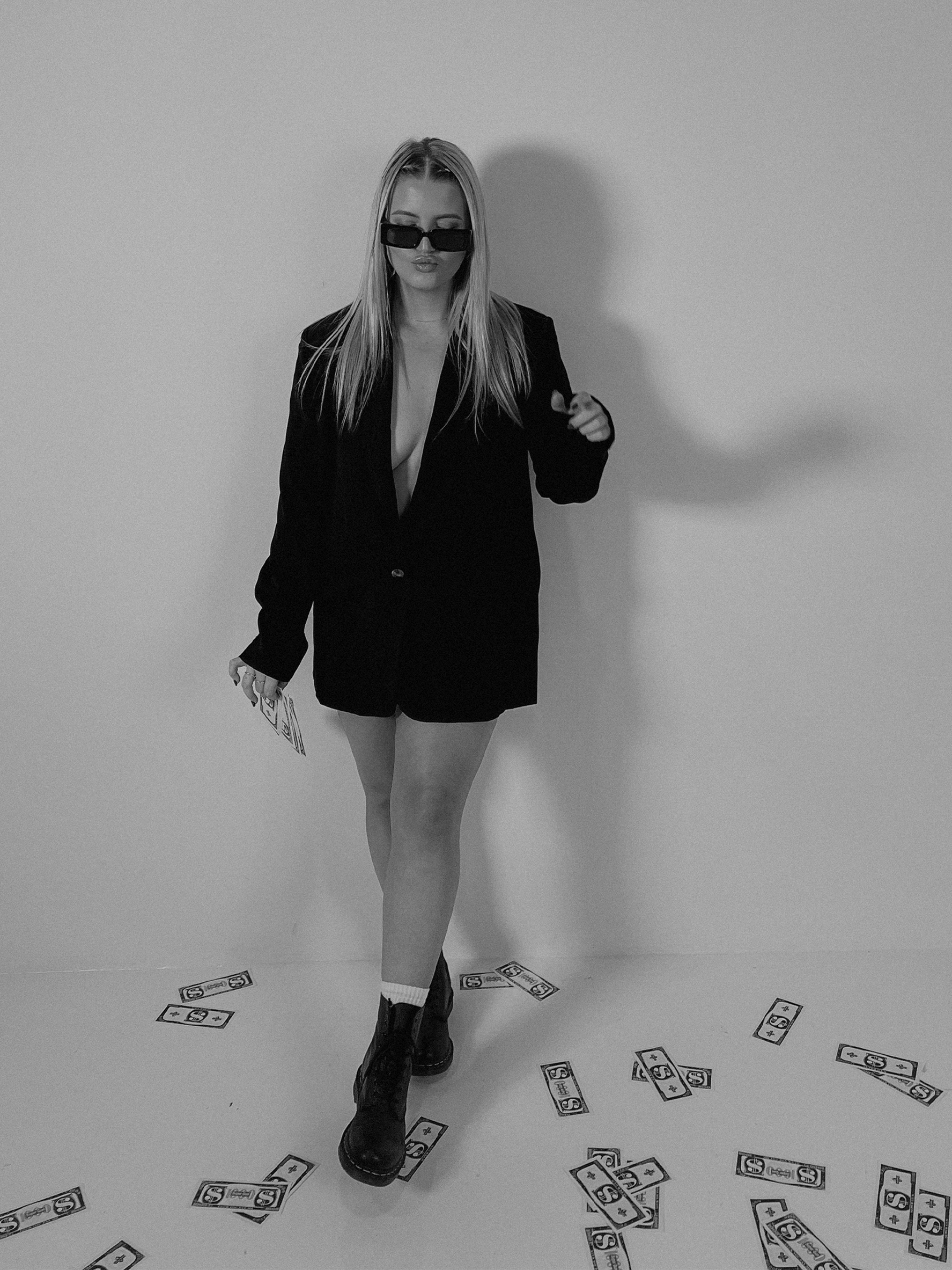 Jess Morgan wearing sunglasses, a black blazer, and chunky boots, standing against a plain wall with scattered fake dollar bills on the floor.