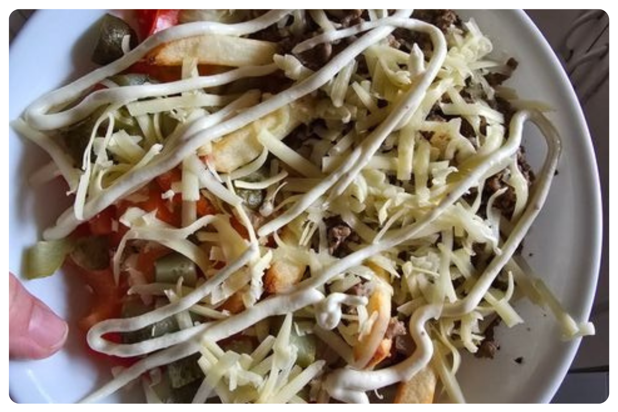 A plate of Mexican taco salad with shredded cheese, ground beef, lettuce, tomatoes, pickles, cucumbers, fried tortilla chips, and topped with sour cream and shredded cheese.
