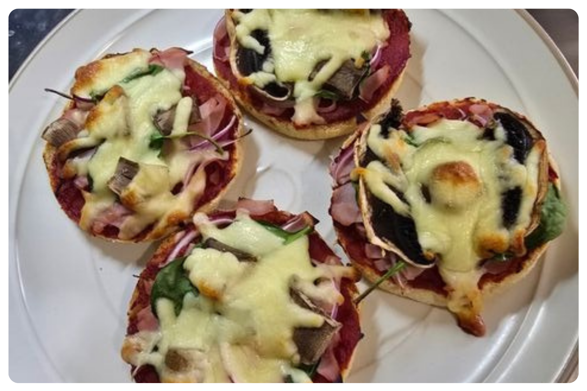 Four mini pizzas with melted cheese, ham, green bell peppers, black olives, and tomato sauce on a white plate.