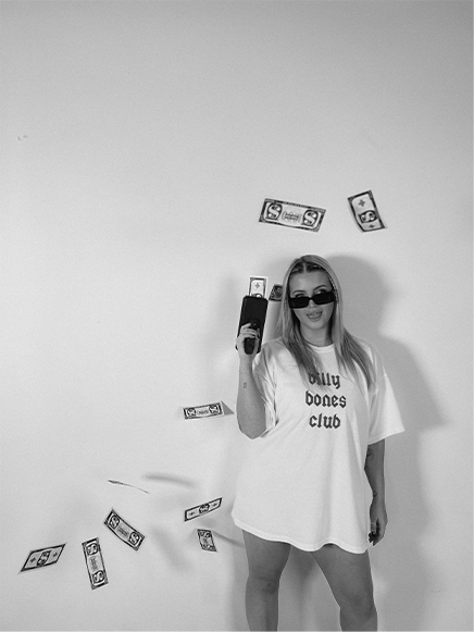 Jess Morgan wearing sunglasses and a large T-shirt with text stands against a plain wall, holding a phone, with fake dollar bills floating around her.