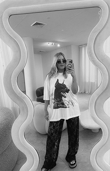 Jess Morgan taking a mirror selfie in a modern, minimalistic room with curved furniture. She wears sunglasses, a white t-shirt with a dog graphic, and lace pants.