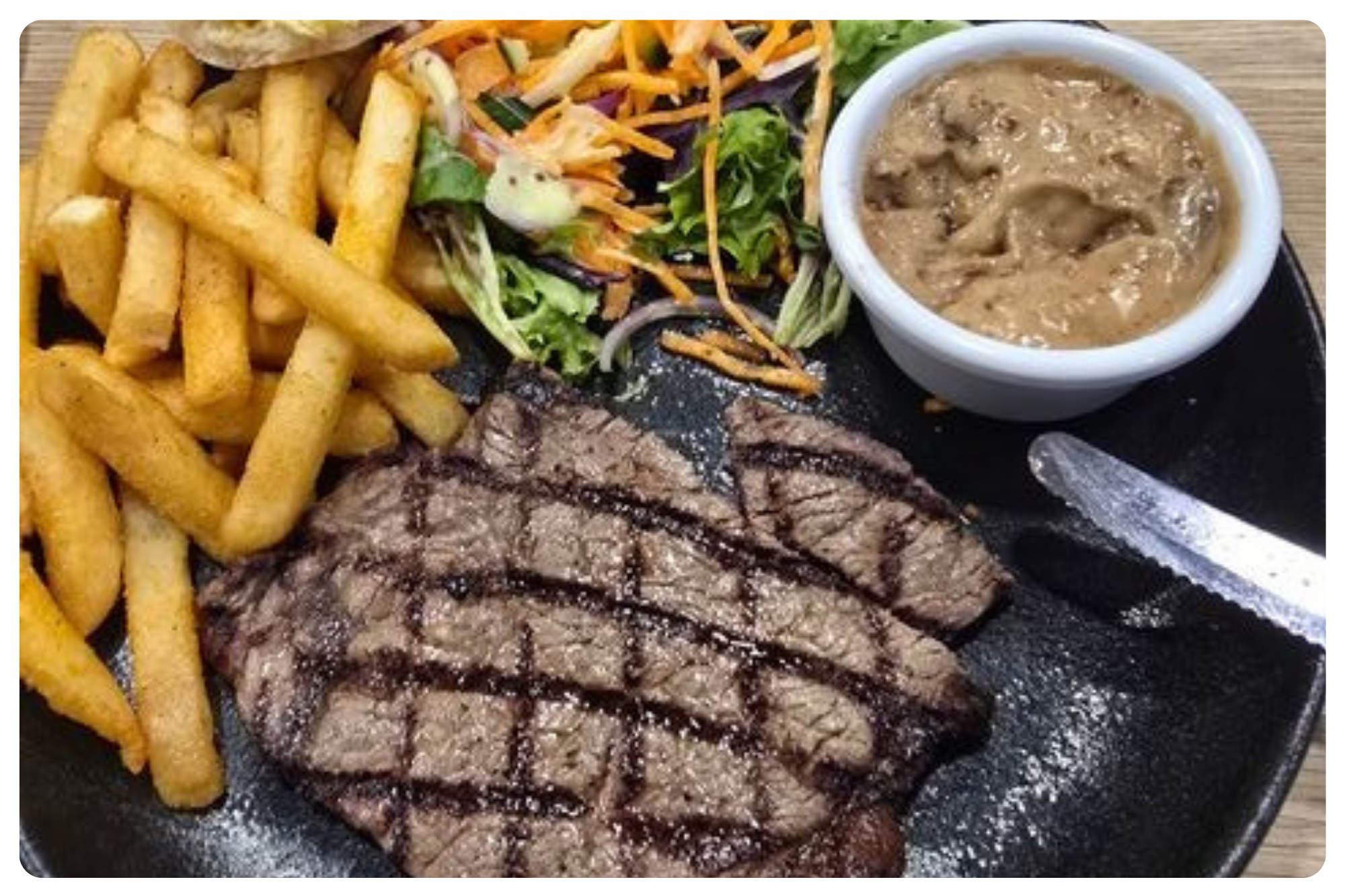 A grilled steak with crisscross grill marks, accompanied by French fries, a small salad, a bowl of gravy, and a fork and knife on a black plate.