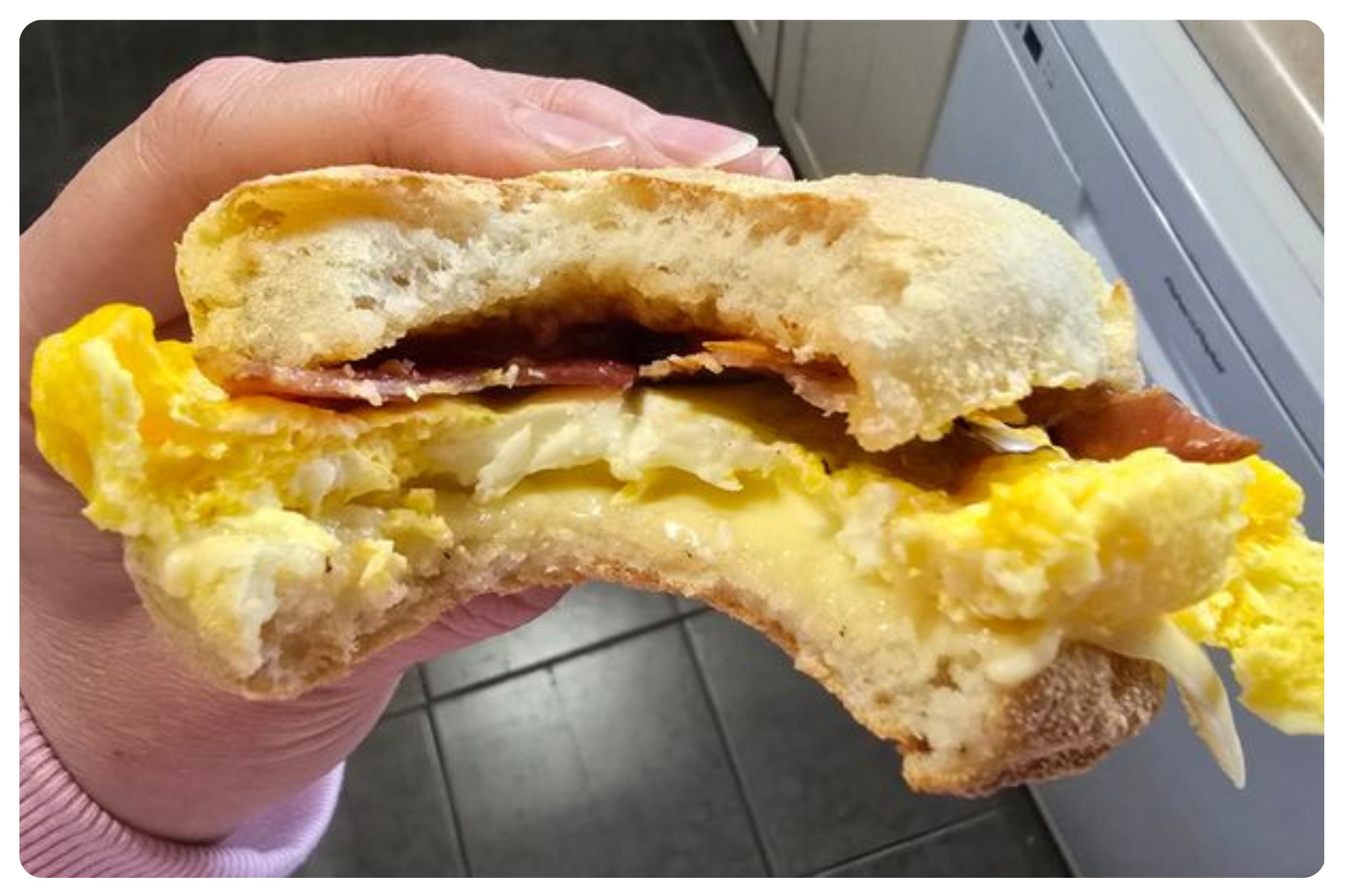 Close-up of a breakfast sandwich with scrambled eggs, cheese, ham, and toasted bread held in a person's hand.