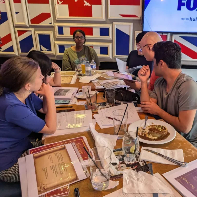 A group of seven people sitting around a wooden table at a restaurant. They are engaged in discussion with papers, menus, and drinks in front of them. The background features a wall with red, white, and blue panels and a TV screen.