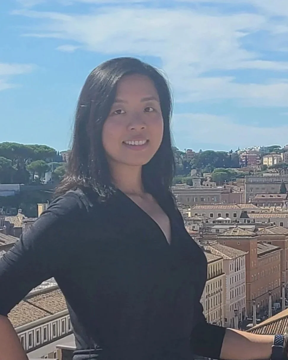 A woman with shoulder-length black hair smiling while standing outdoors on a sunny day with a cityscape in the background.