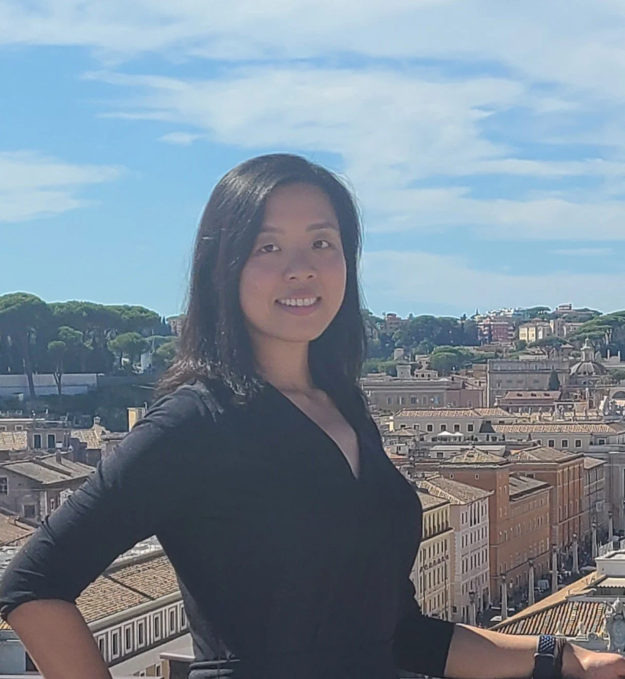 A woman with black shoulder-length hair wearing a black top, smiling, with a cityscape and blue sky in the background.