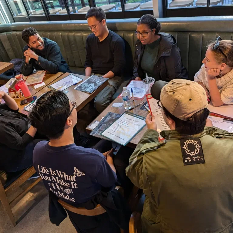 Group of seven young adults sitting around a table in a restaurant, engaged in conversation and looking at menus.