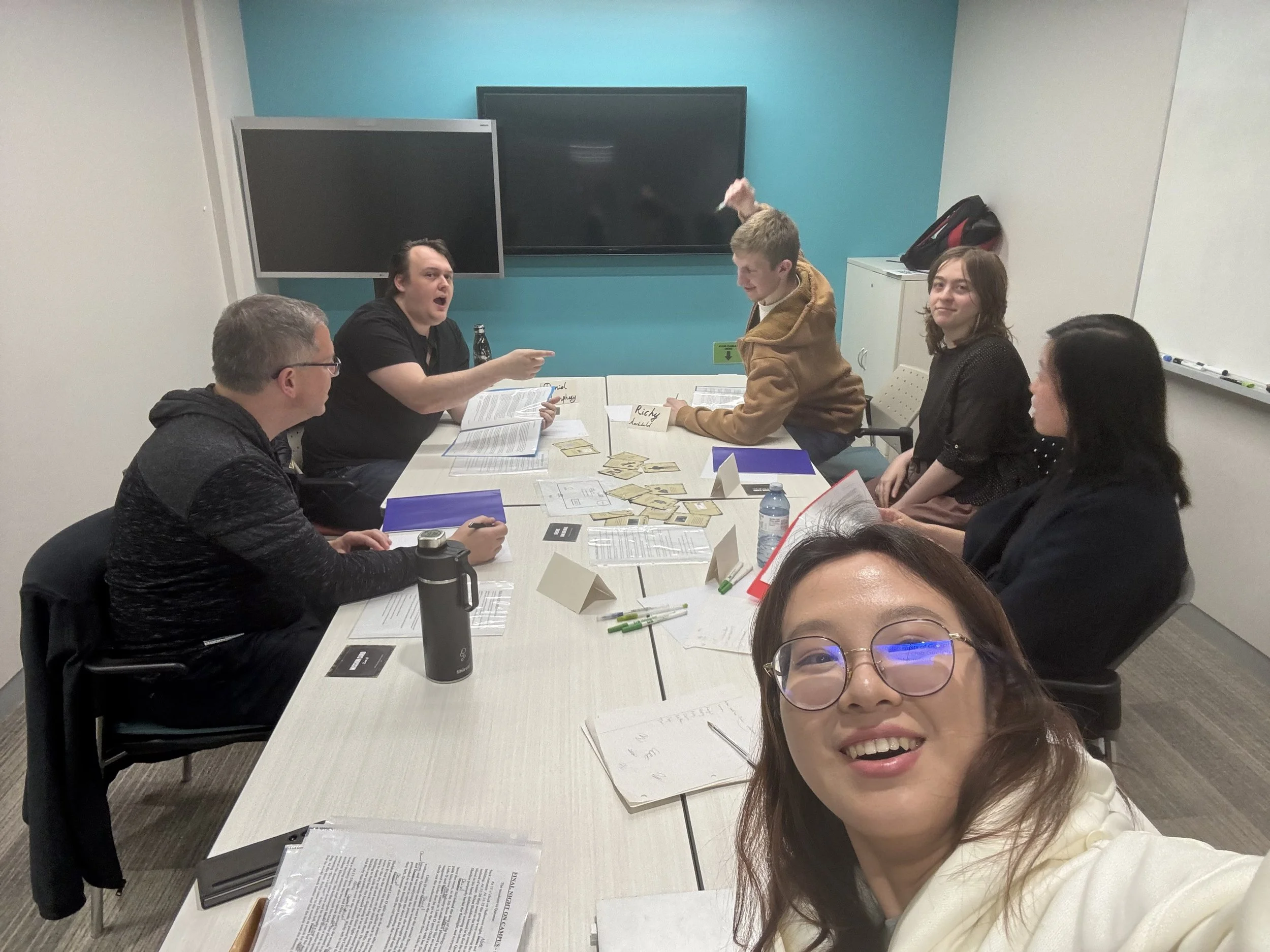 Group of five people sitting around a conference table, appearing to be playing a tabletop role-playing game with character sheets, cards, and pens, in a brightly colored office or conference room.