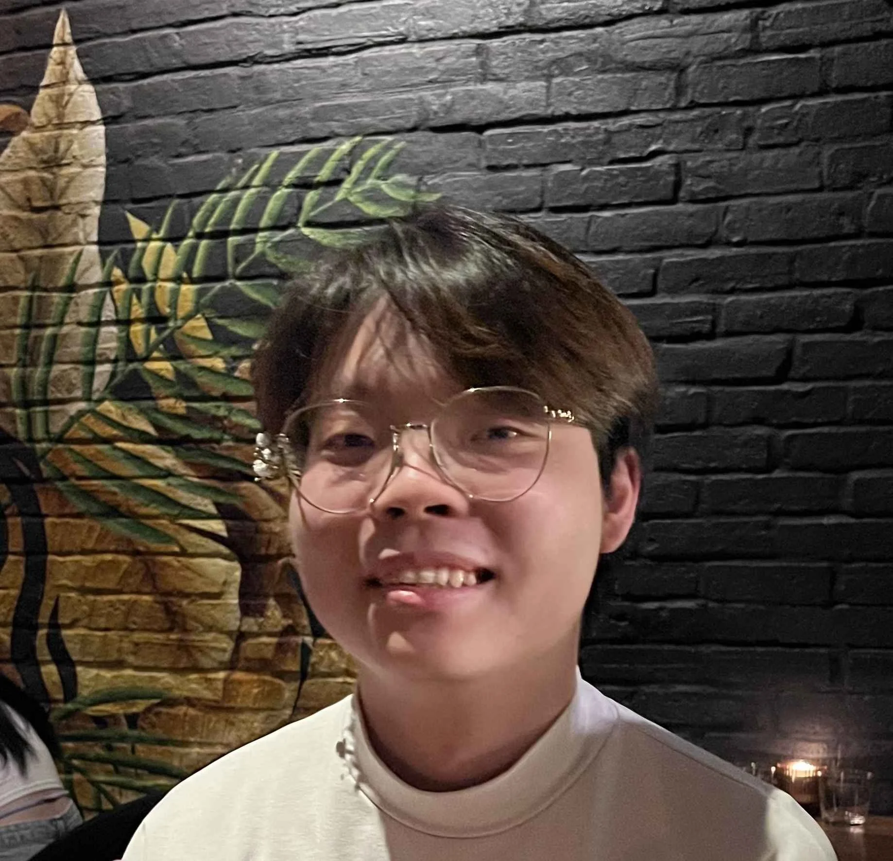 A young person with short brown hair and glasses, smiling in front of a black brick wall with a painted leaf design.