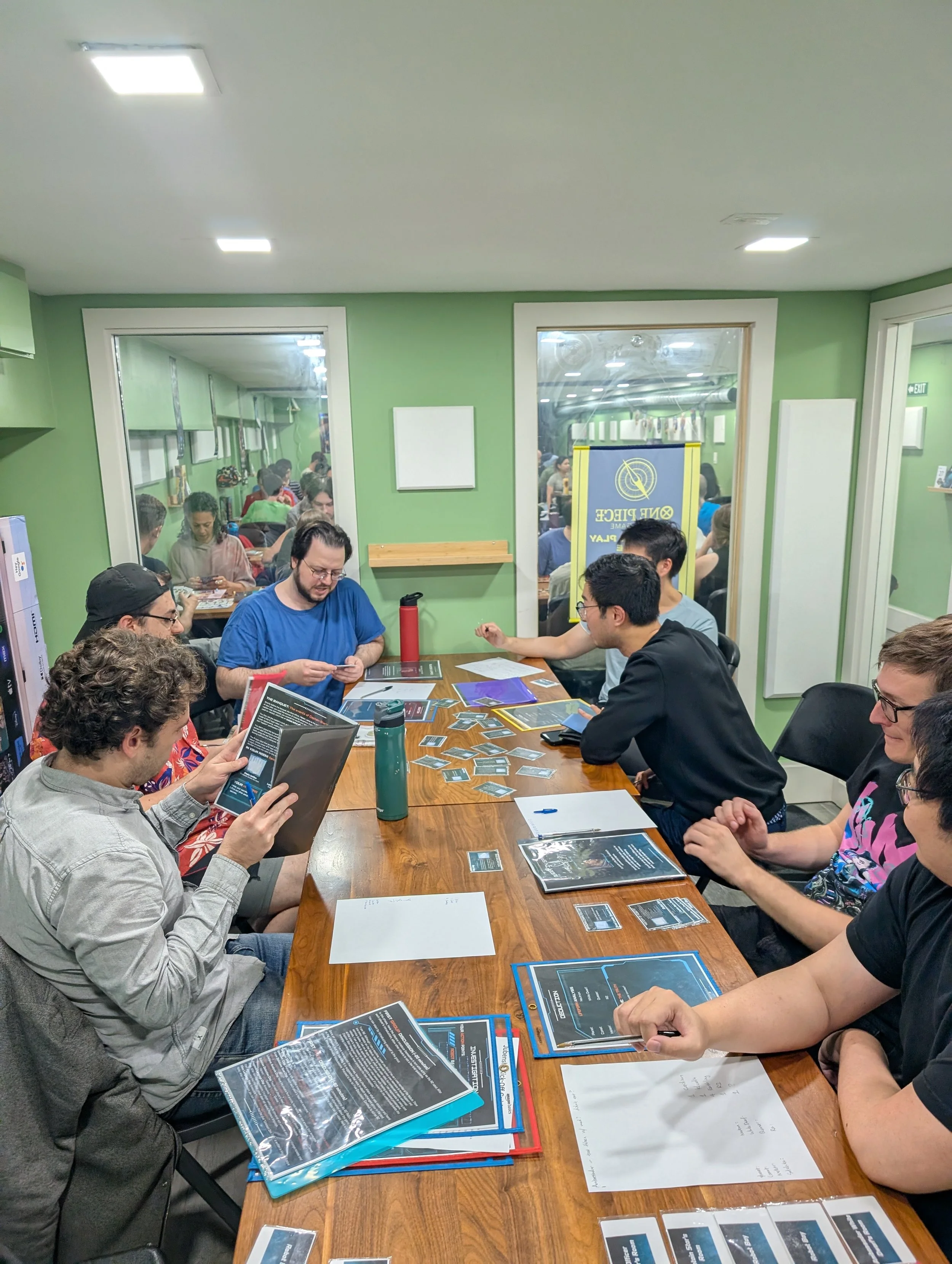 People playing a tabletop card game at a wooden table in a room with green walls and windows showing an adjacent room with more people gaming.