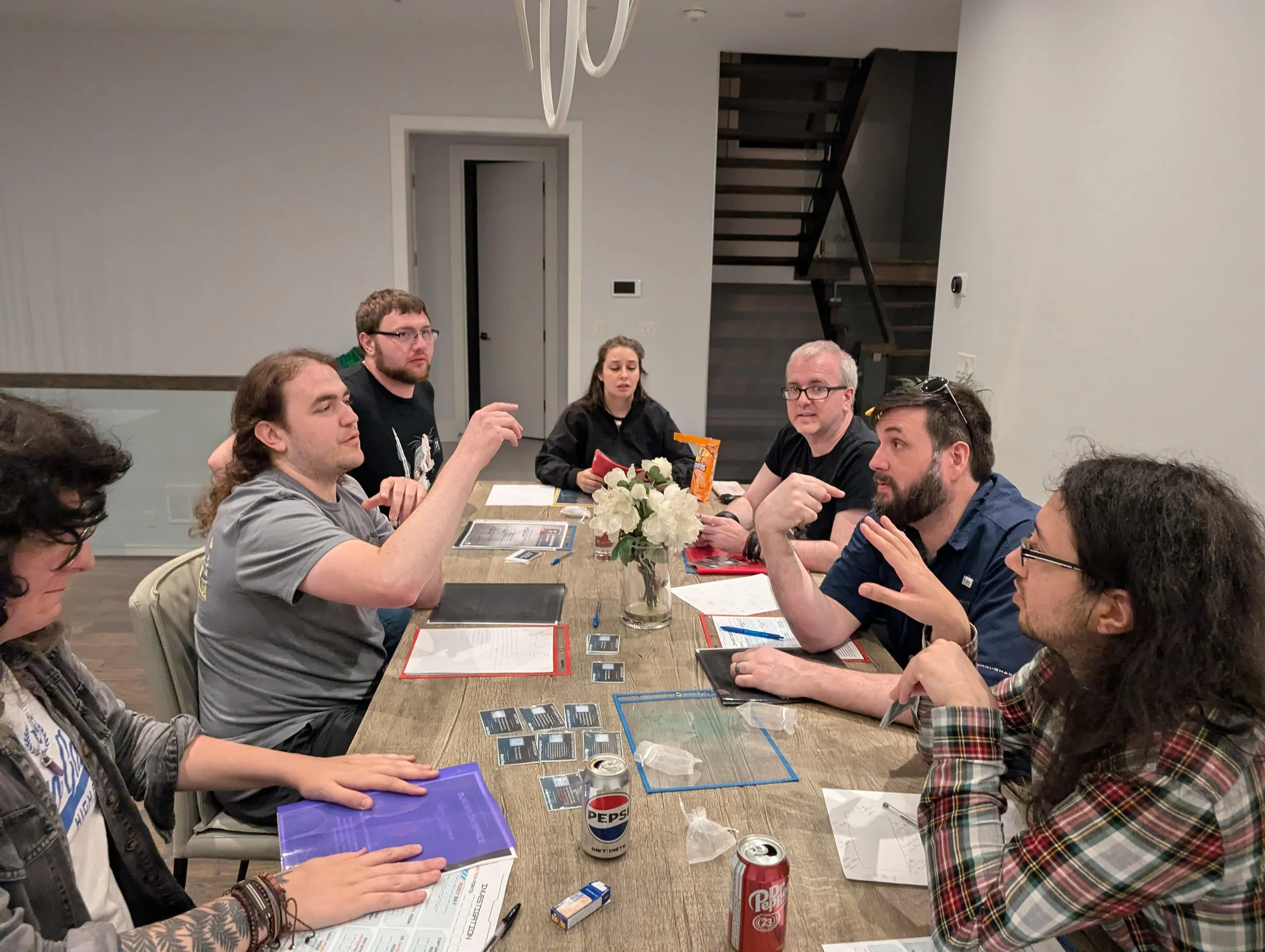 Group of seven people sitting around a wooden table engaged in a board game, with game cards, notebooks, and drinks on the table in a modern indoor setting.