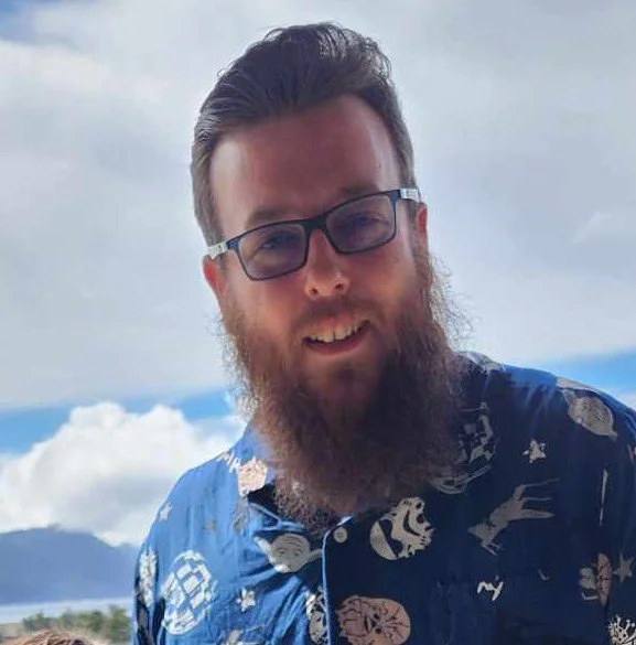 A man with glasses and a long beard smiling outdoors, with clouds and mountains in the background.