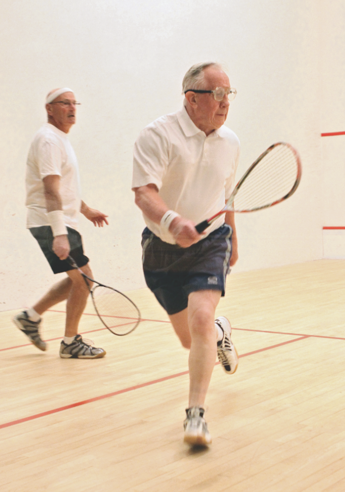 Two older men playing squash on an indoor court, both holding squash racquets and wearing athletic clothes.