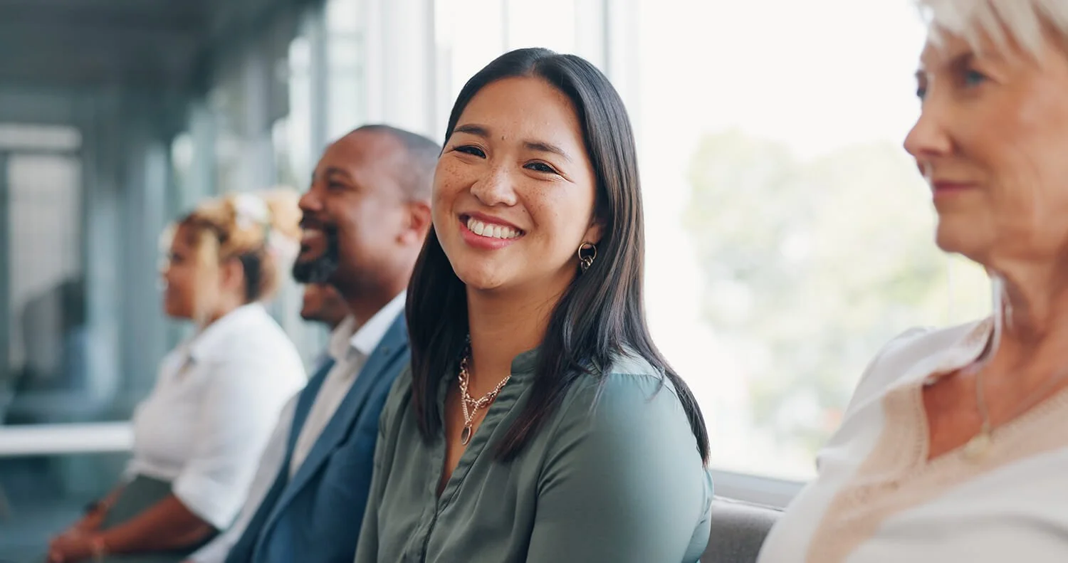 A diverse group of people sitting and smiling in a well-lit waiting area or conference room, making use of the AI Validation Studio, to help test a business solution using AI.