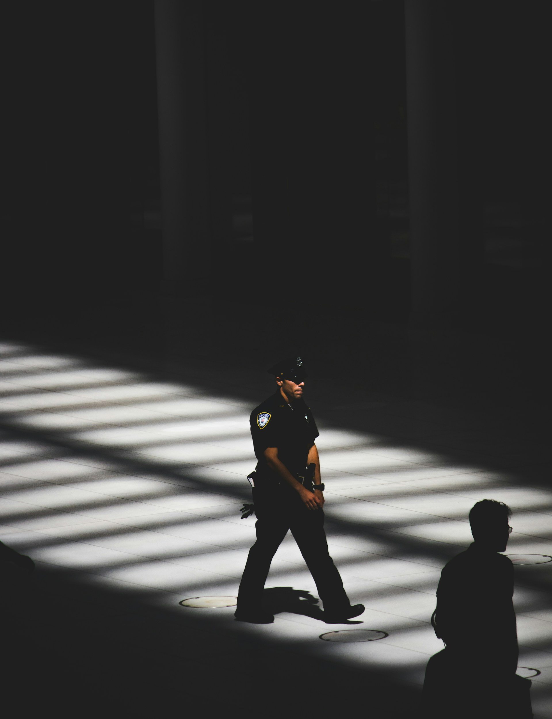 A police officer walking on a illuminated grid floor with shadow stripes, with another person sitting nearby, in a dark space.