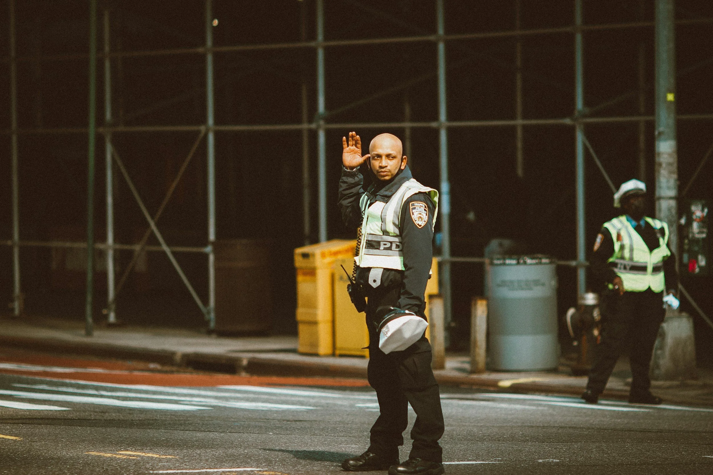 A police officer standing in the street at night, waving with his right hand. He is wearing a reflective safety vest over a black uniform, with a badge on his arm. Another officer is in the background, standing near a scaffold and trash cans.