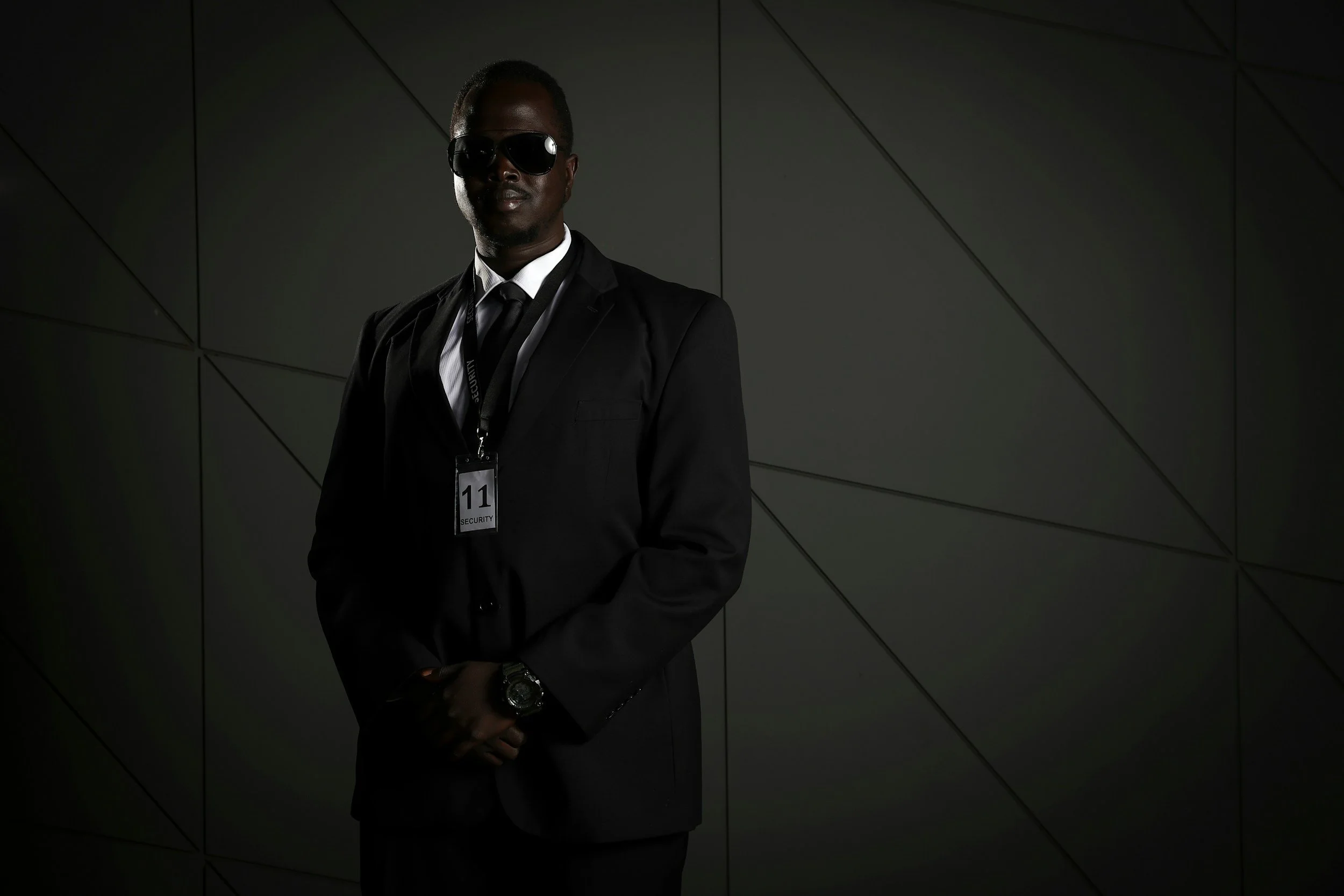Security guard in black suit and sunglasses standing against a dark geometric wall, wearing a security badge with the number 11.