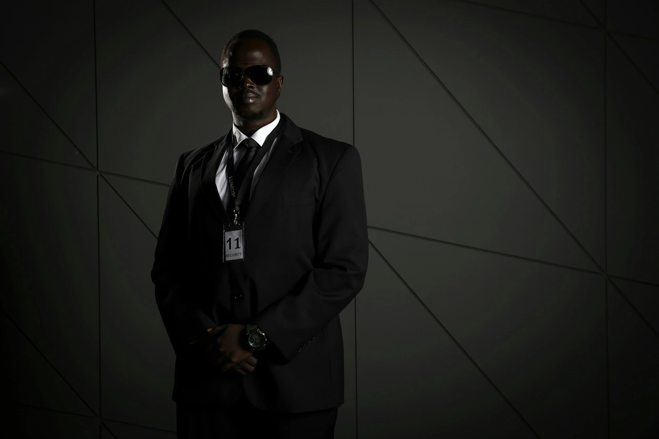 Man in black suit with sunglasses, security badge, and watch standing against a dark, geometric wall background.