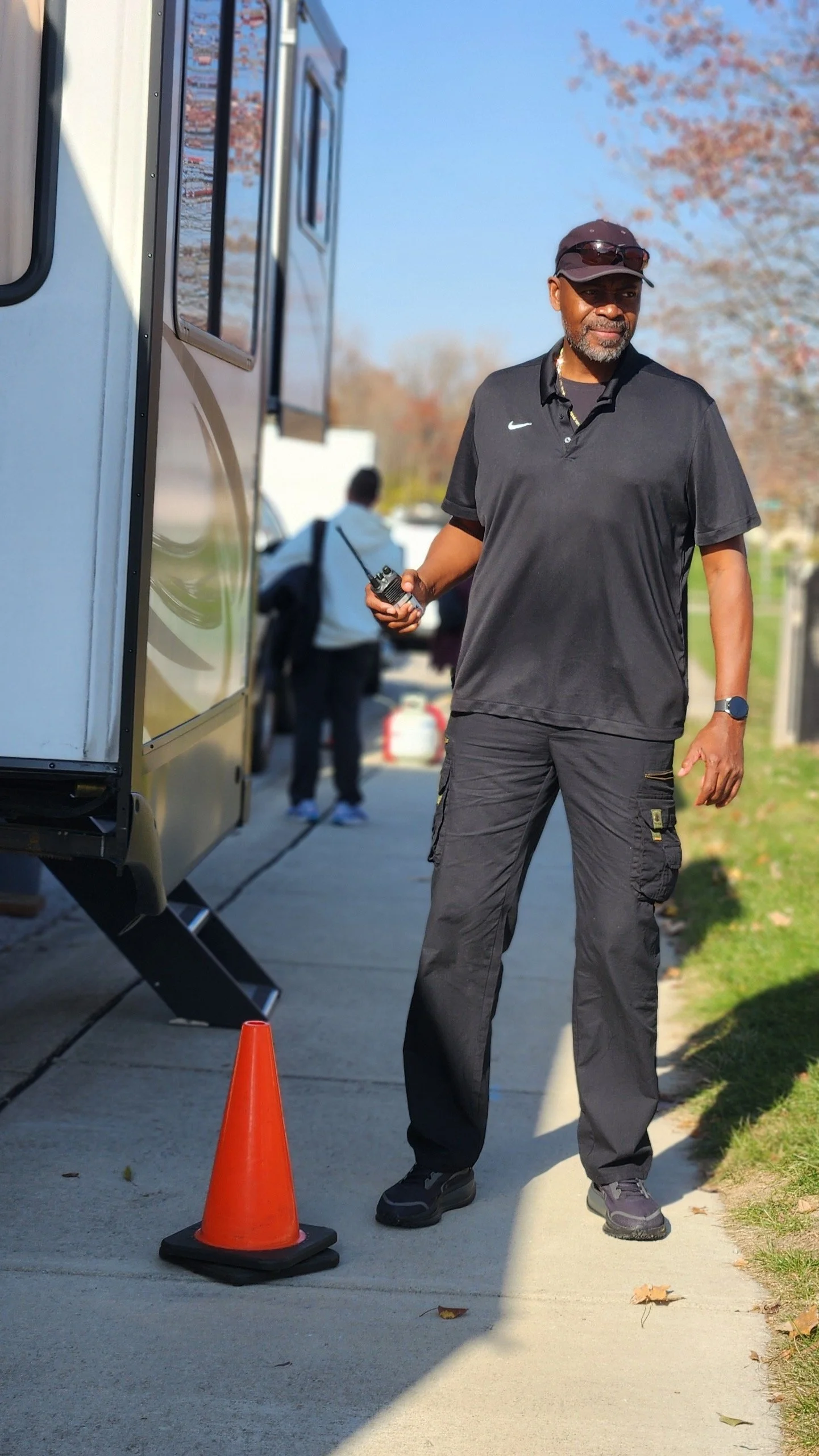 A man dressed in black standing next to a white mobile food truck, holding a walkie-talkie, with an orange traffic cone on the sidewalk. There are blurred people in the background and trees with fall foliage.