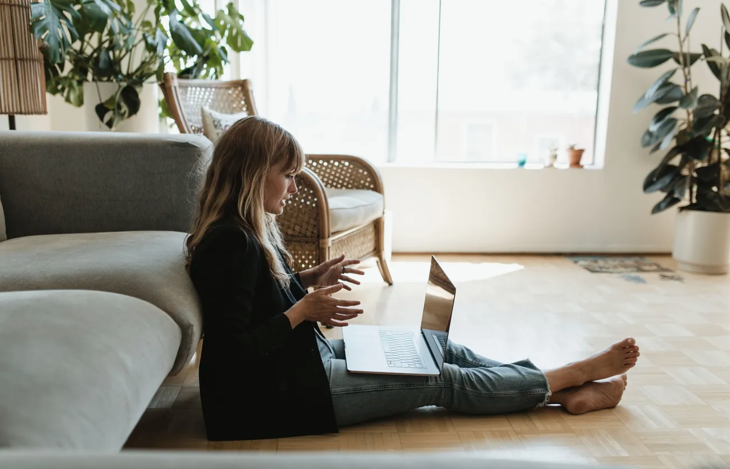 A woman sitting on the floor with a laptop, gesturing with her hands in a bright living room with large windows, plants, and rattan furniture.