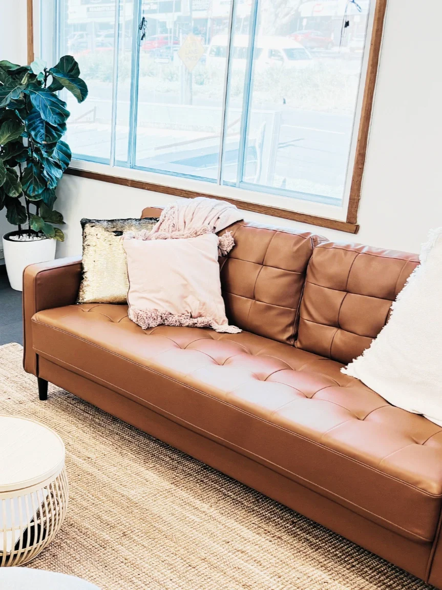 A brown leather couch with three cushions in pink, beige, and gold, placed next to a large window with a view of parked cars outside. A green potted plant is beside the couch, and a woven rug is underneath.
