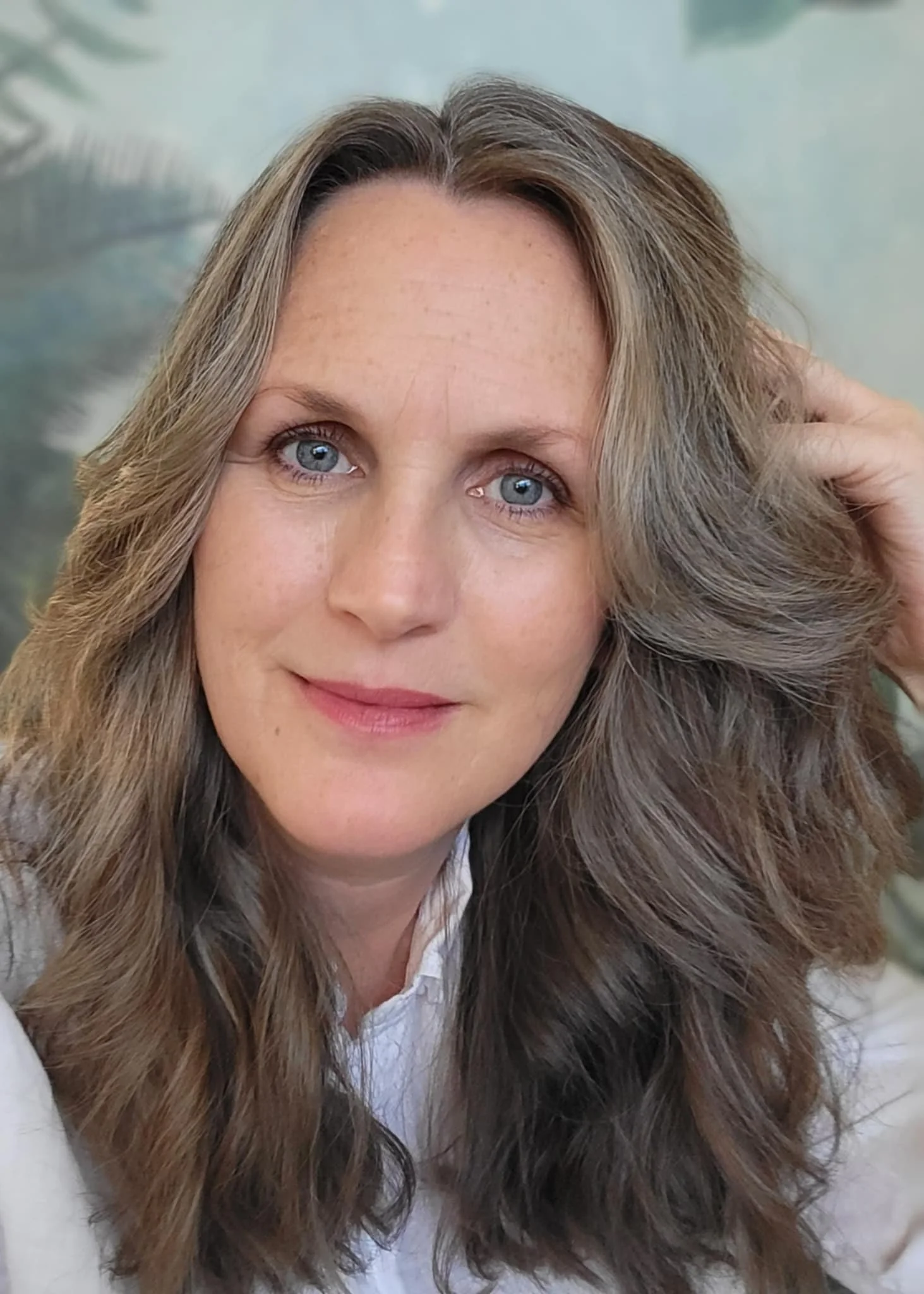 A close-up portrait of a woman with wavy light brown hair and blue eyes, smiling softly, wearing a white shirt with a blurred outdoor background.