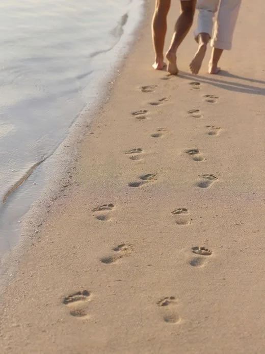 Two people walking barefoot on a sandy beach, leaving footprints in the sand.
